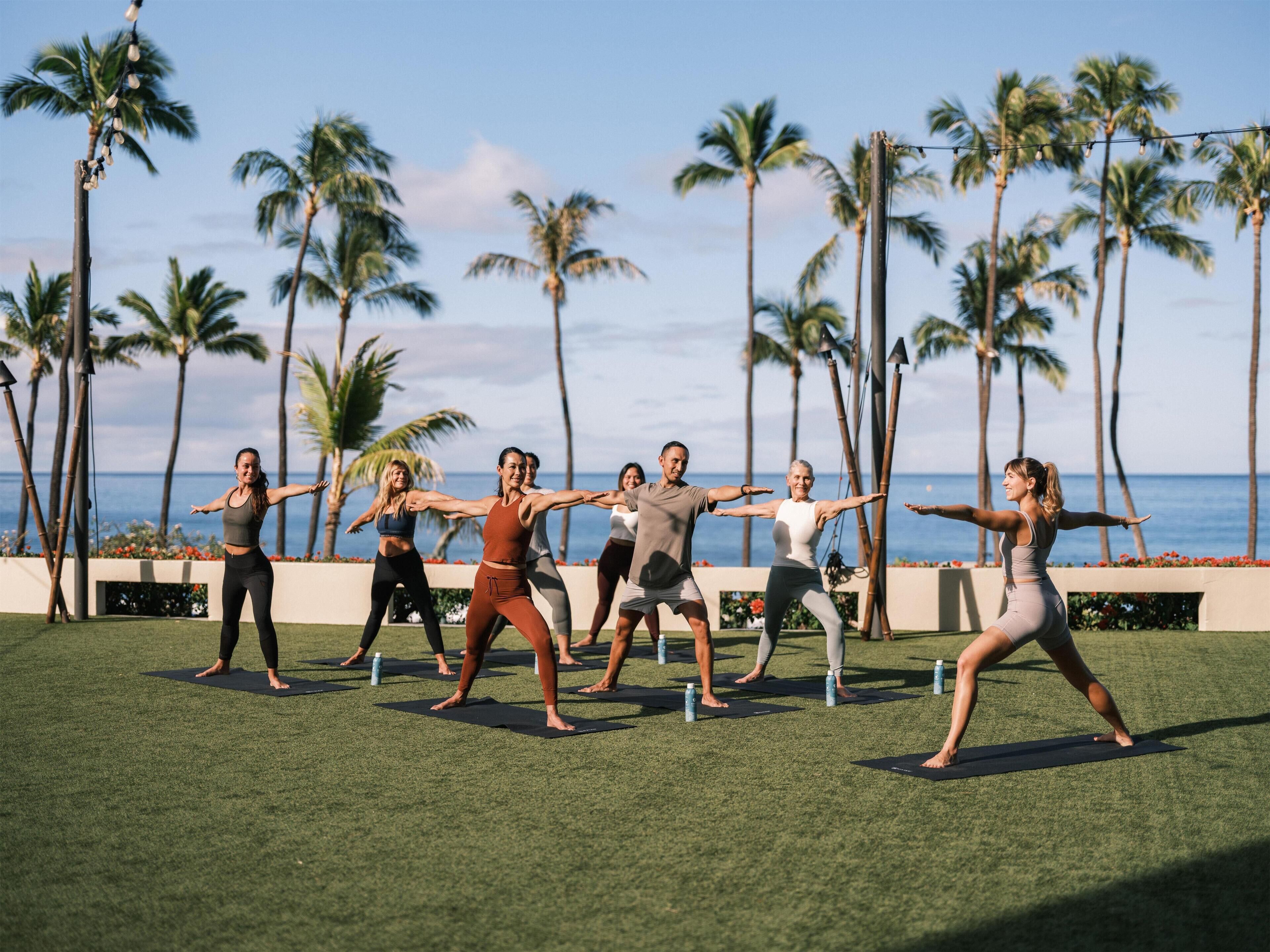 Un grupo de personas realizando ejercicios de yoga al aire libre, con vista a palmeras y al océano de fondo.