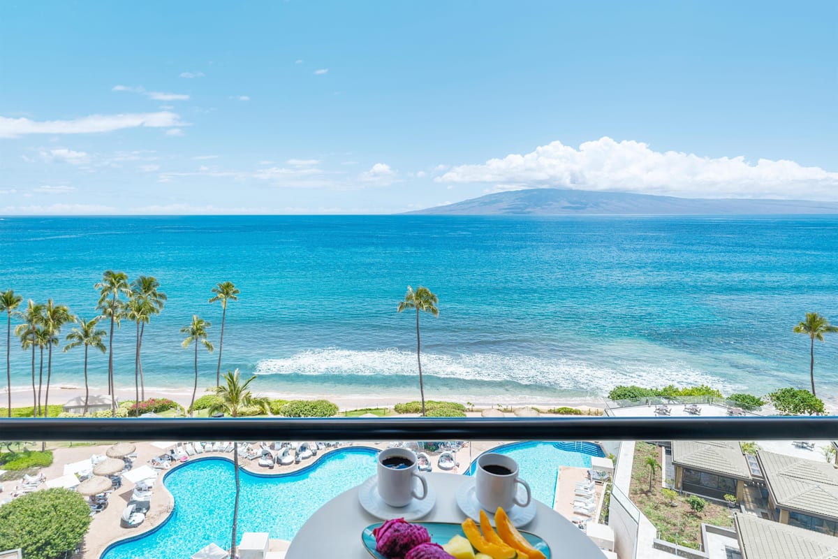 A poolside view with a clear ocean and beach, a mountain in the distance, and a dining area with a table and chairs.