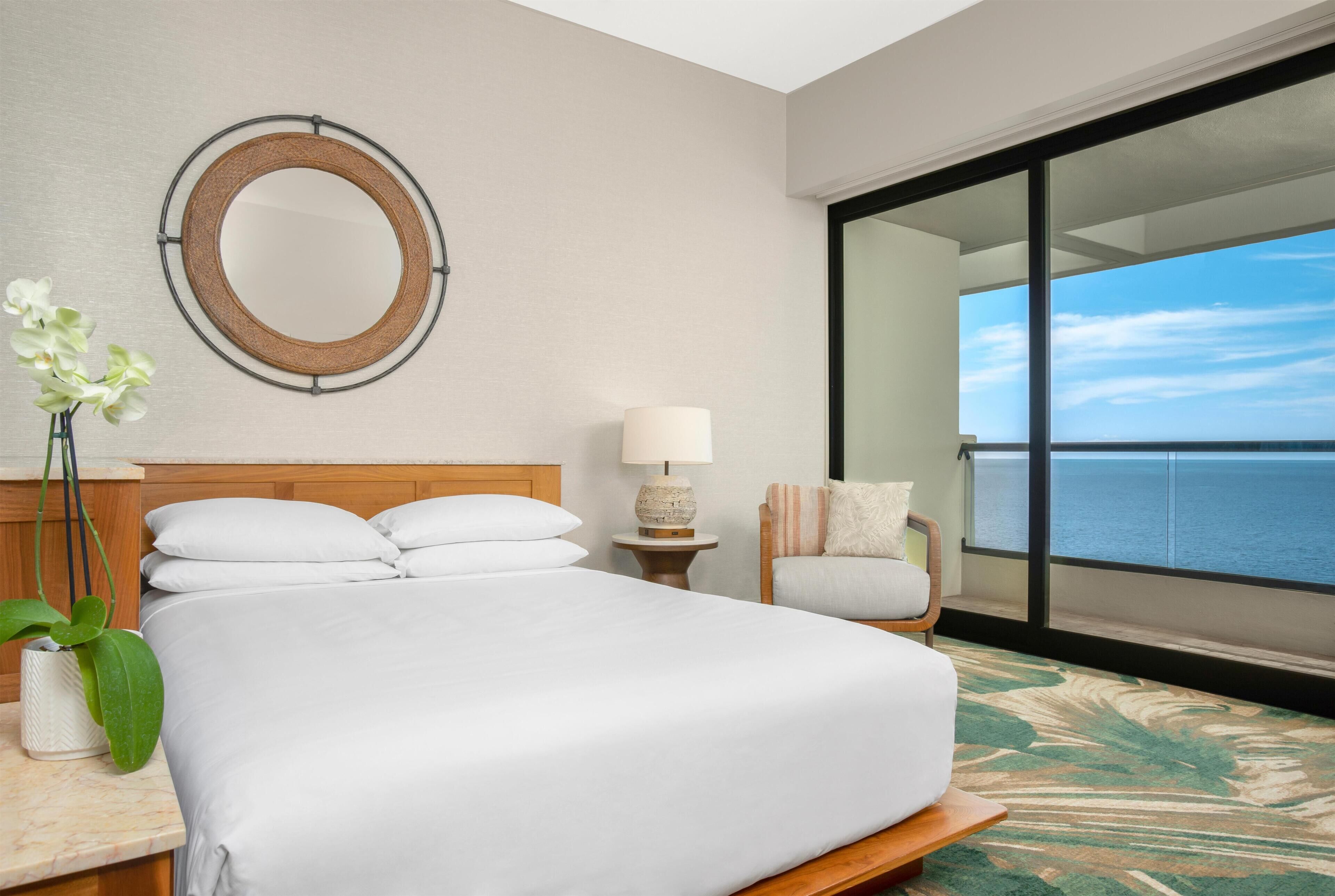 A hotel room with a bed, a round mirror, a nightstand with a lamp, a chair, and a view of the ocean.