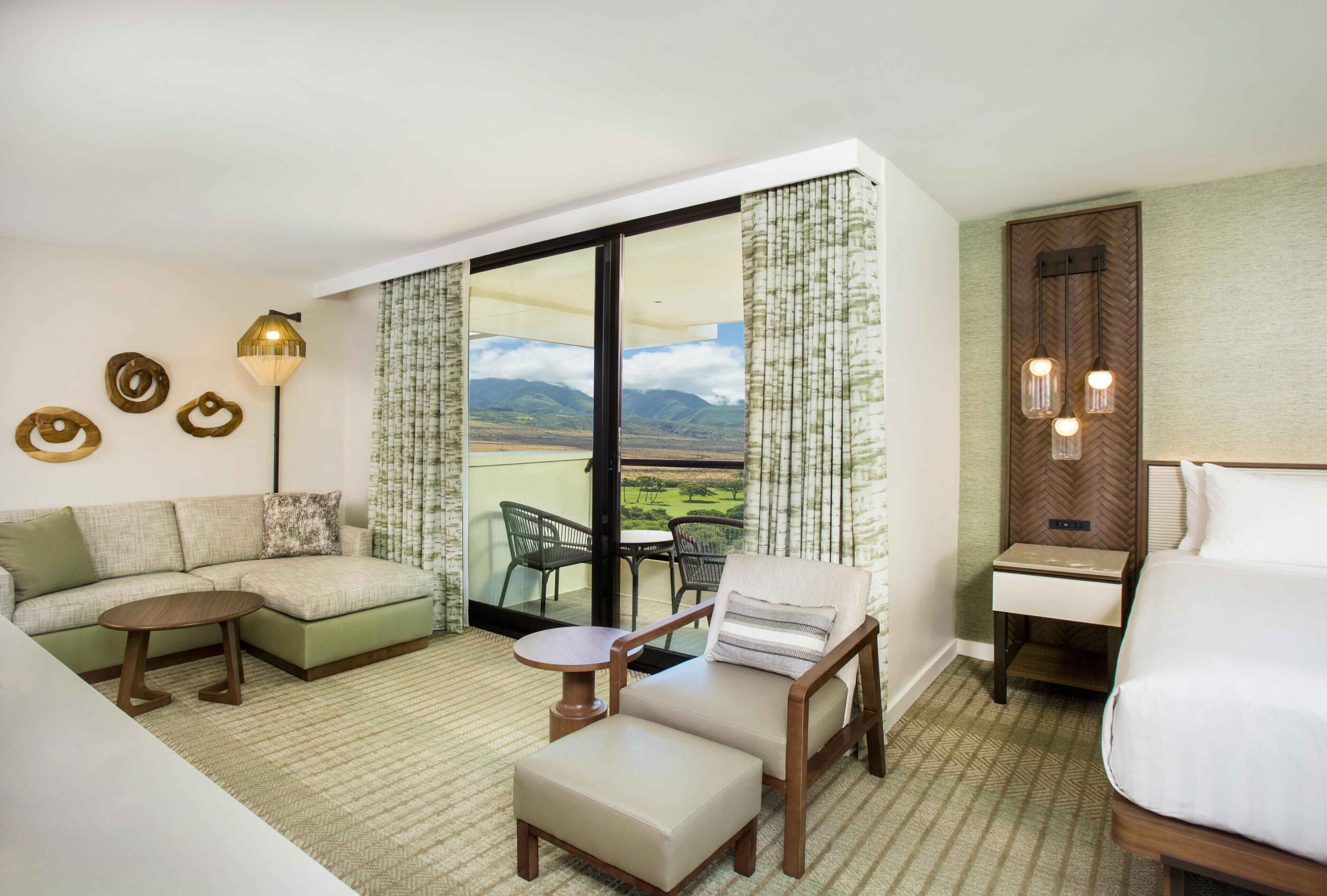Habitación de hotel con sofá, una mesita, una silla, cama y balcón con vistas a las montañas.