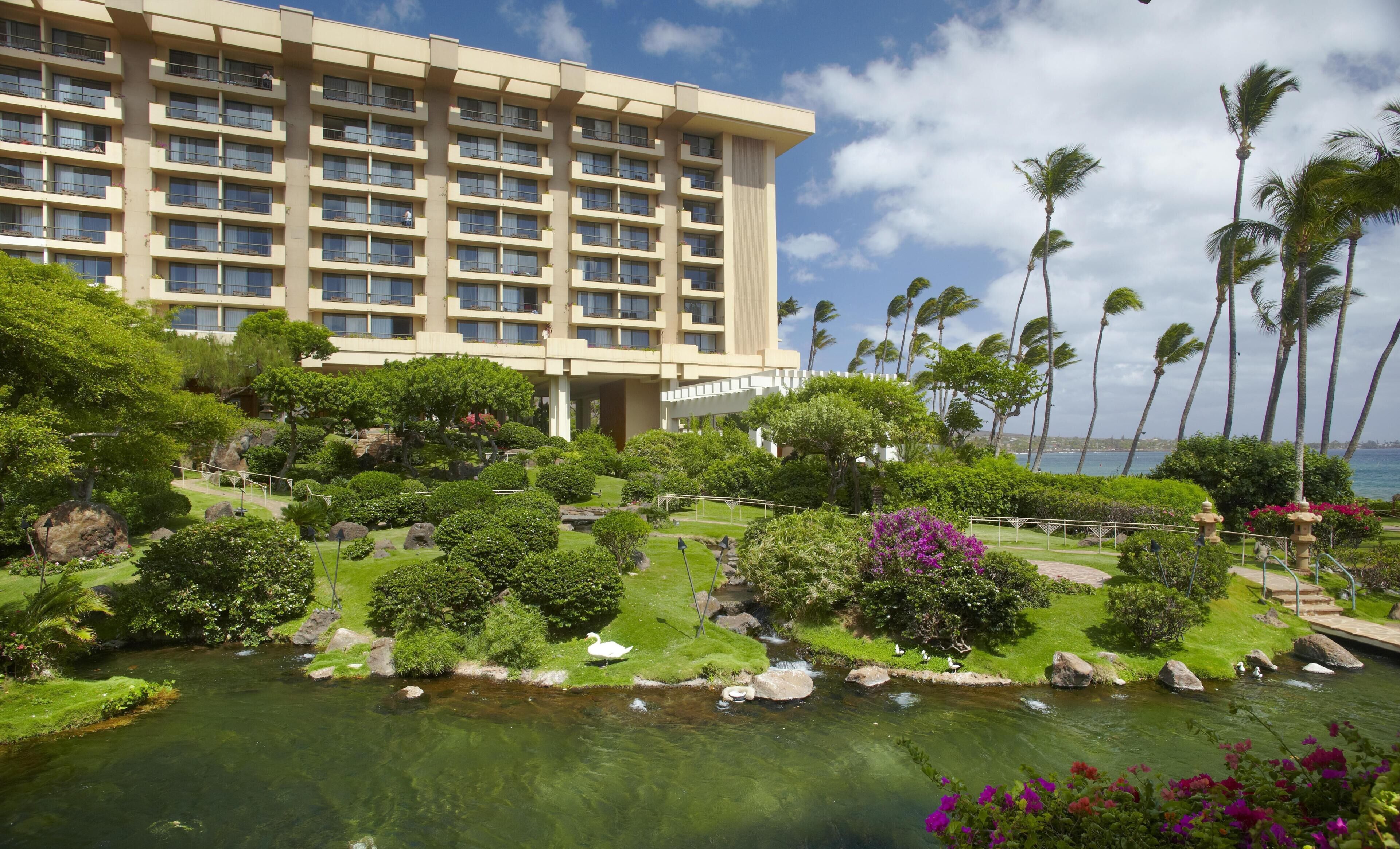 A hotel with a large building, a landscaped garden with a pond, and palm trees.