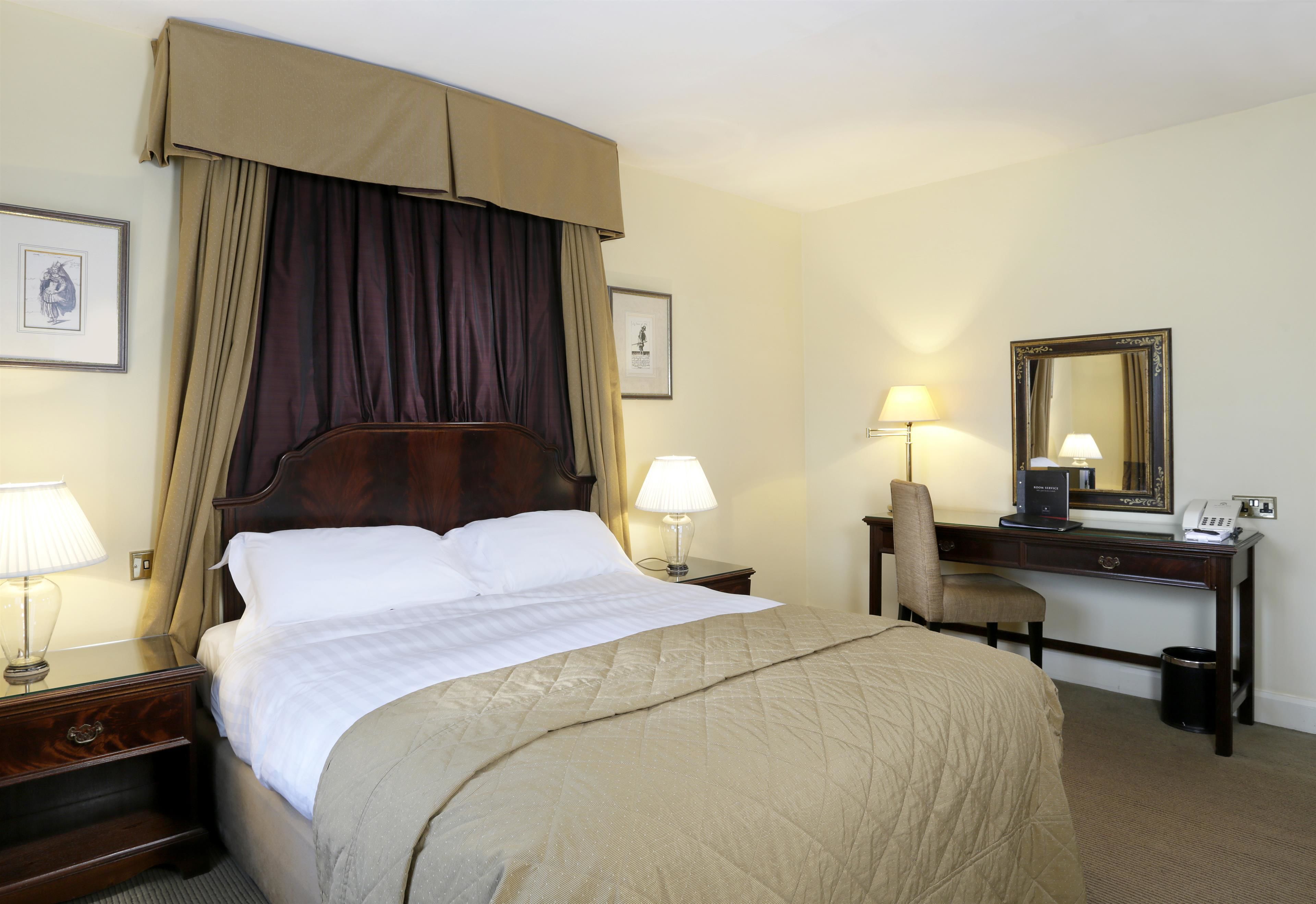 A hotel room with a large bed, two bedside tables with lamps, a desk with a chair, a mirror, and a picture on the wall.