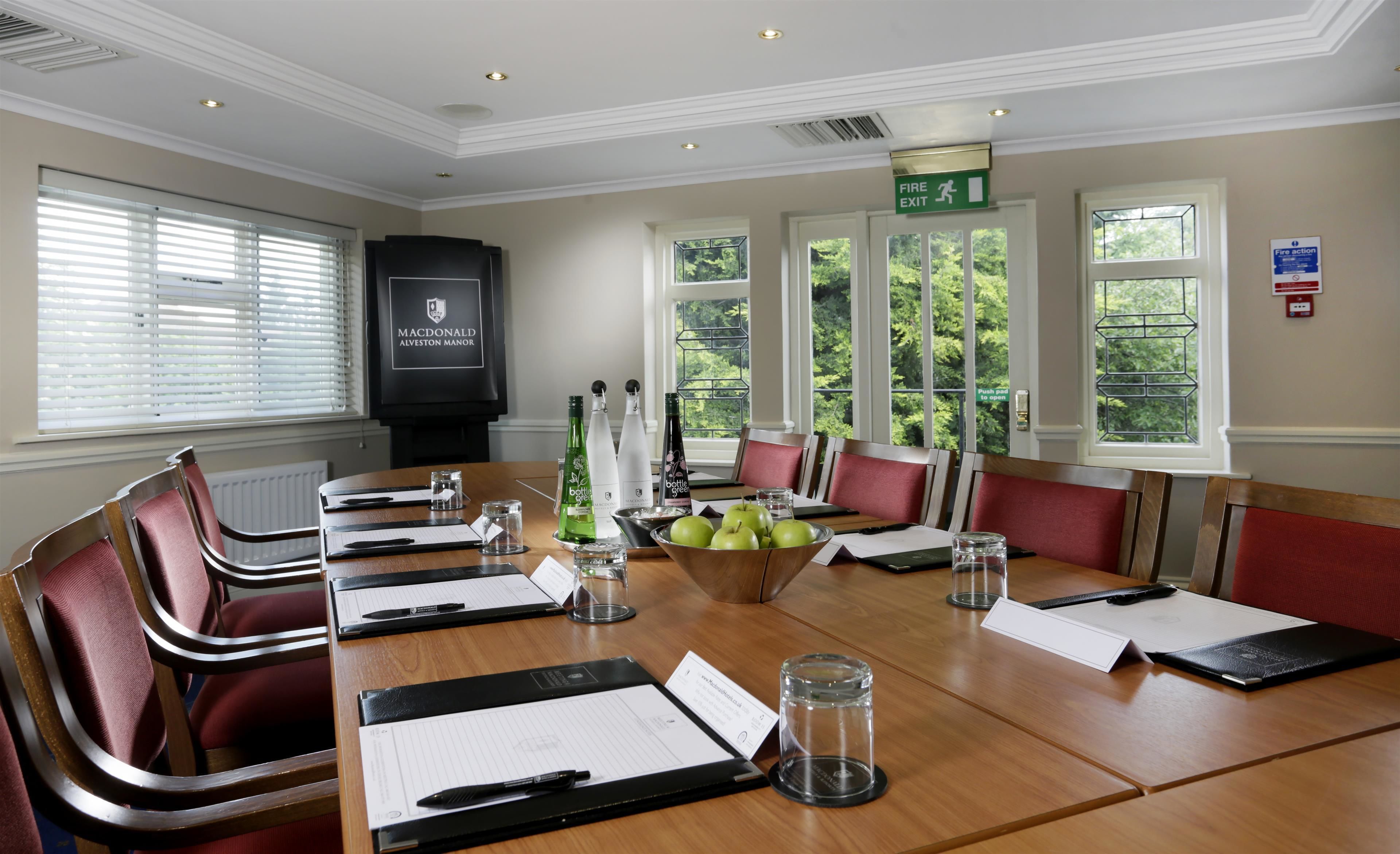 A conference room with a long table, red upholstered chairs, and a large screen displaying the hotel's name.