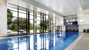 An indoor swimming pool with clear glass walls, a tiled floor, and a seating area with chairs.