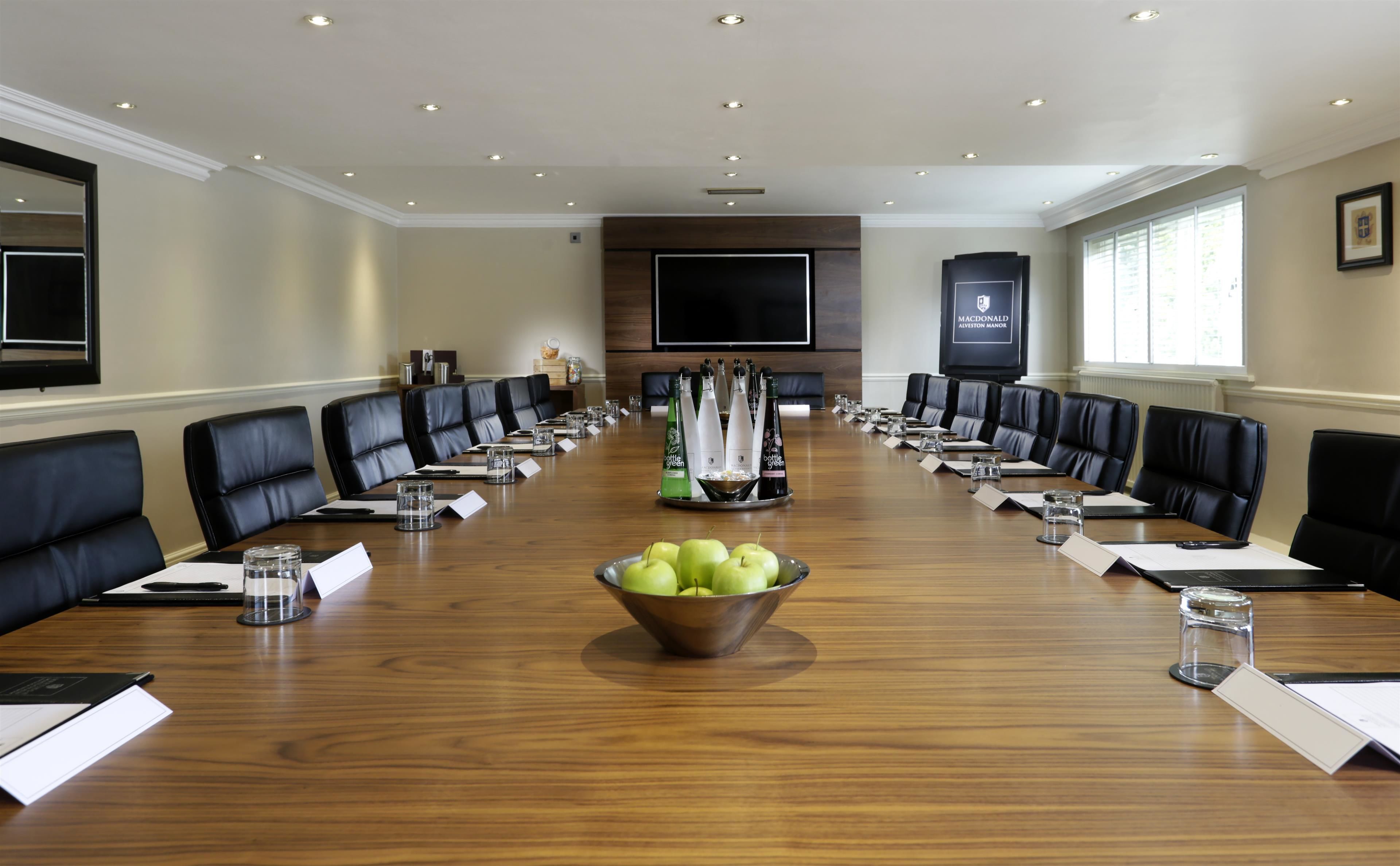 Un salón de conferencias con una mesa larga, sillas de cuero negro, un bol de manzanas y una pantalla de televisión.