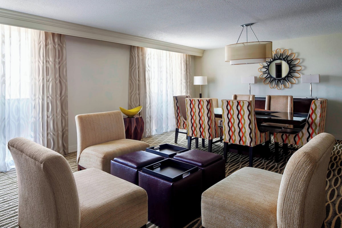 A hotel room with a seating area featuring two armchairs, a sofa, and a coffee table. There is a dining table with chairs and a decorative mirror on the wall.