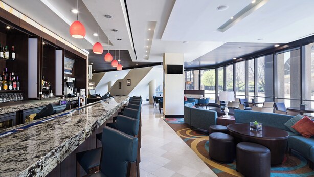 A modern bar area with a granite countertop, bar stools, and a variety of liquor bottles. There is a seating area with sofas and round tables, and large windows providing a view of the outdoors.