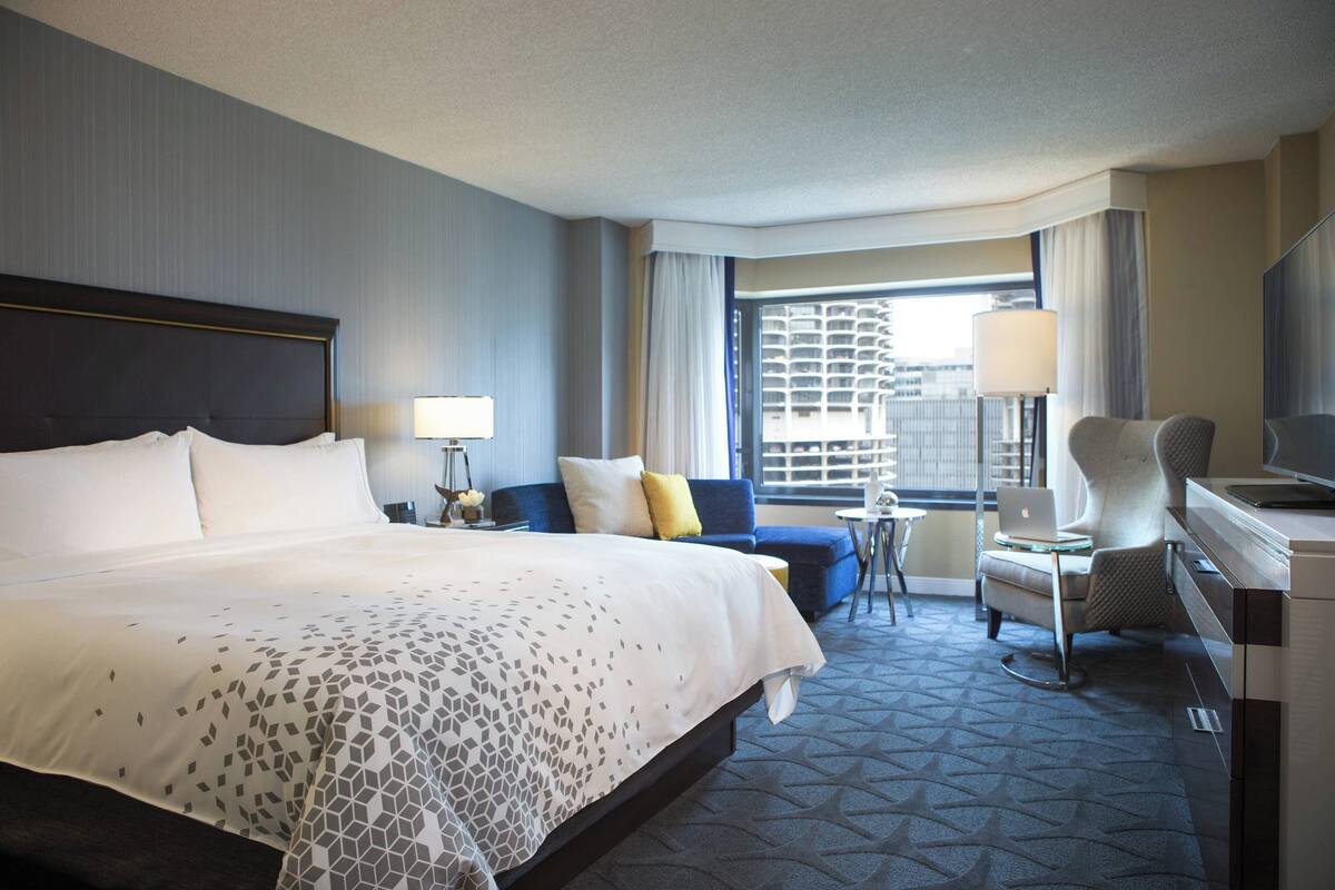 Une chambre d’hôtel avec un grand lit, un bureau avec un ordinateur portable, une chaise, un canapé et une fenêtre avec des rideaux.