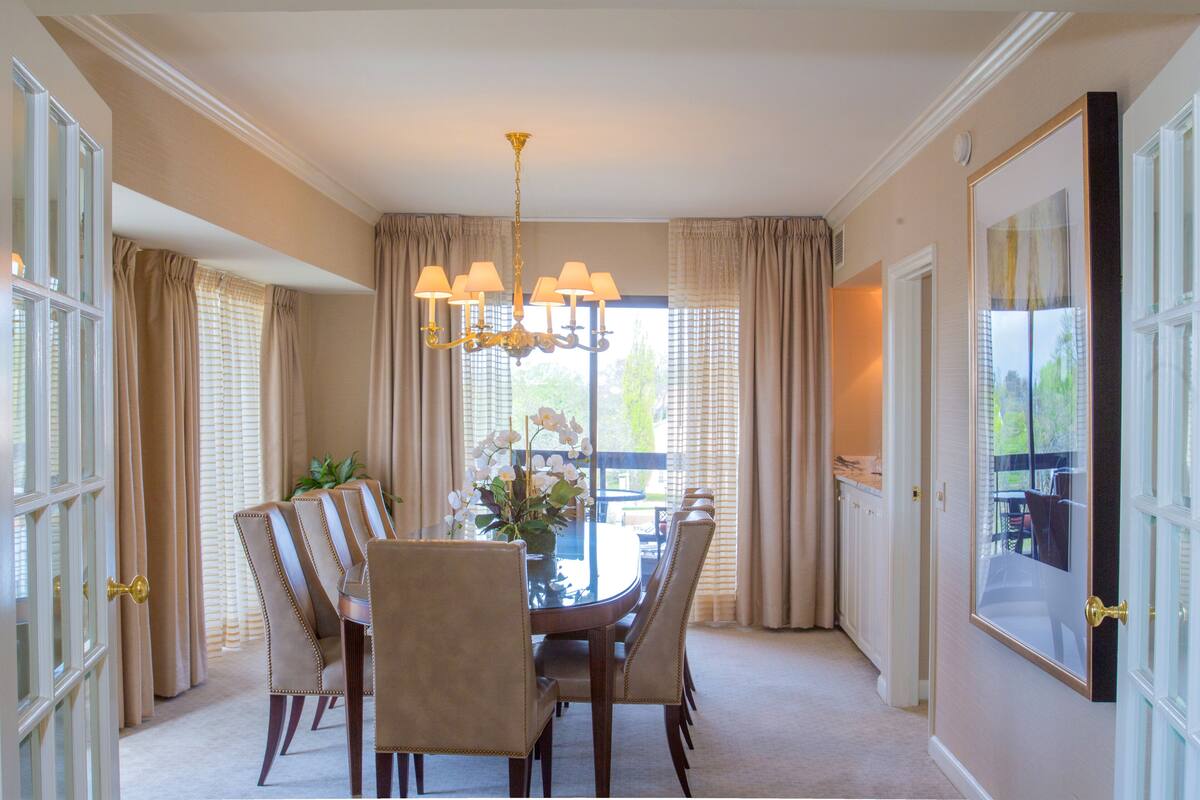 Uma sala de jantar com uma mesa redonda, seis cadeiras, um candelabro e cortinas.