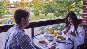 A dining table with breakfast items and drinks, a view of a park, and a building in the background.