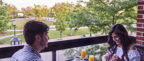 A dining table with breakfast items and drinks, a view of a park, and a building in the background.