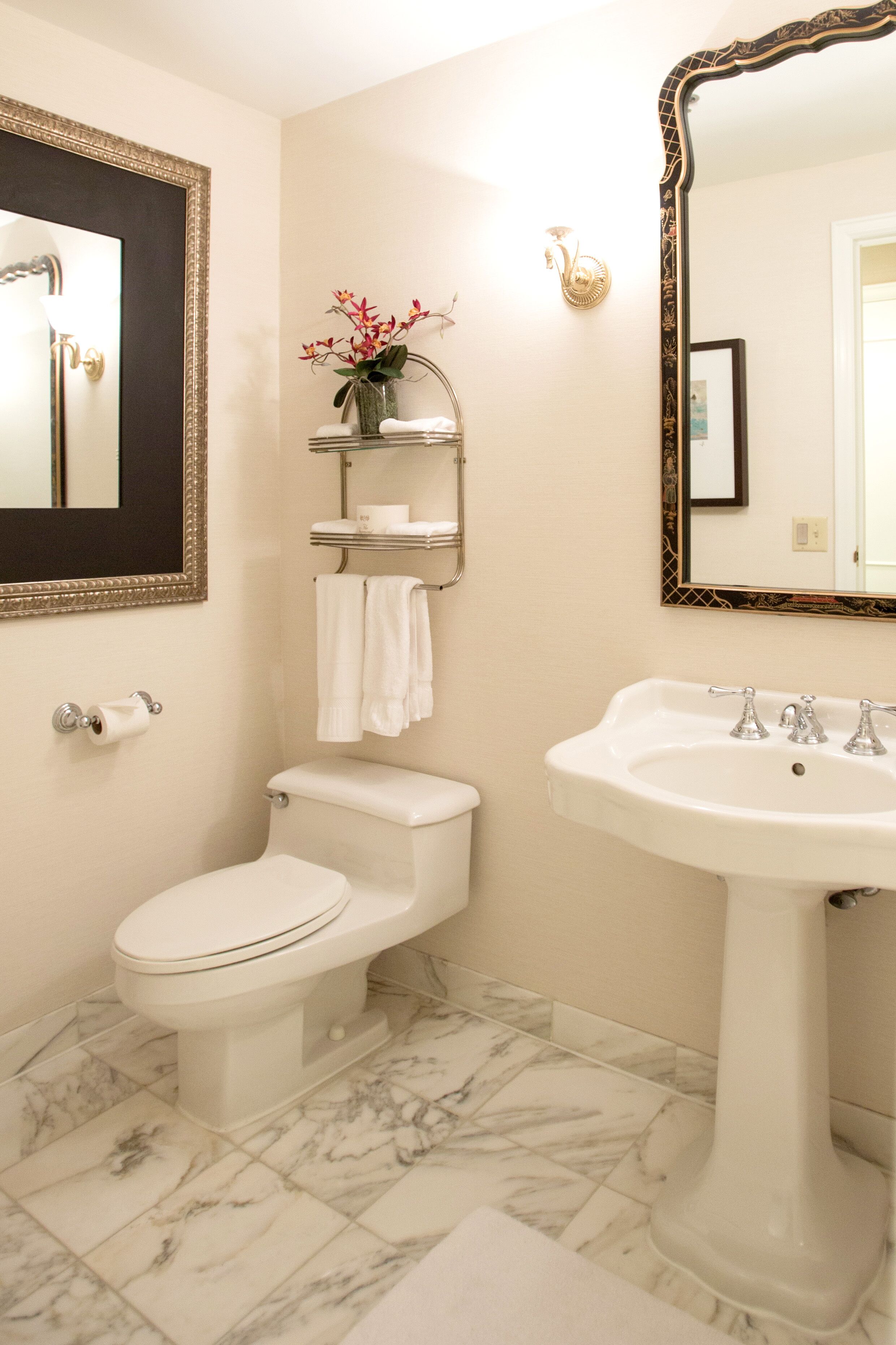 A bathroom with a toilet, sink, and two mirrors. There is a towel rack with a towel and a vase with flowers.