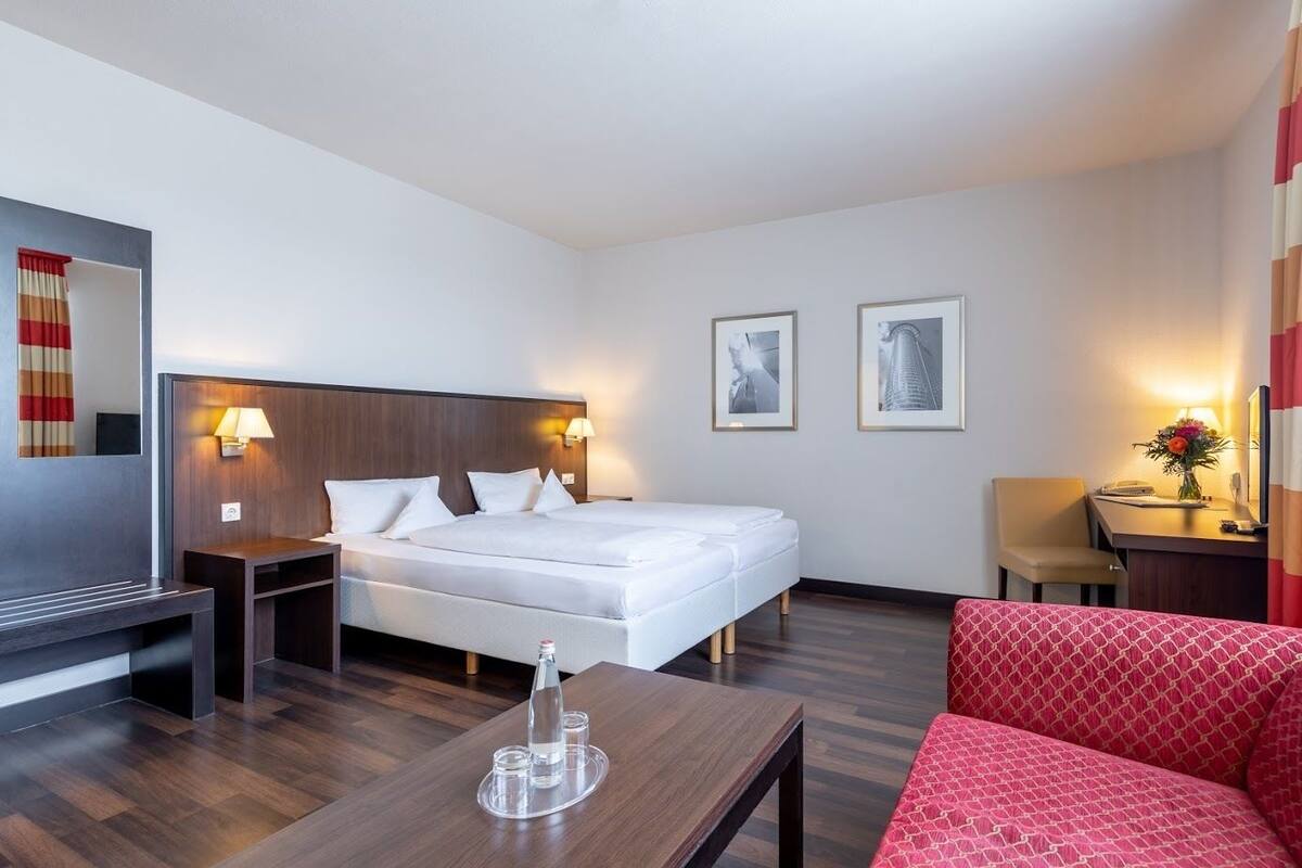 A hotel room with a bed, bedside tables, a desk, a chair, and a red sofa.