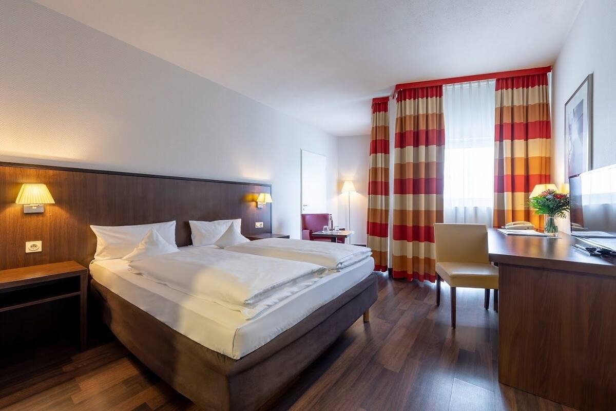 A hotel room with a large bed, two bedside tables with lamps, a desk with a chair, and a large window with striped curtains.