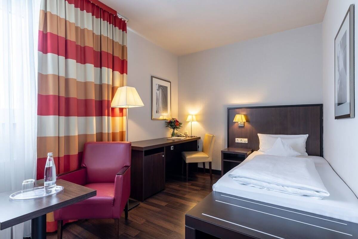 A hotel room with a bed, desk, chair, and a large window with curtains.