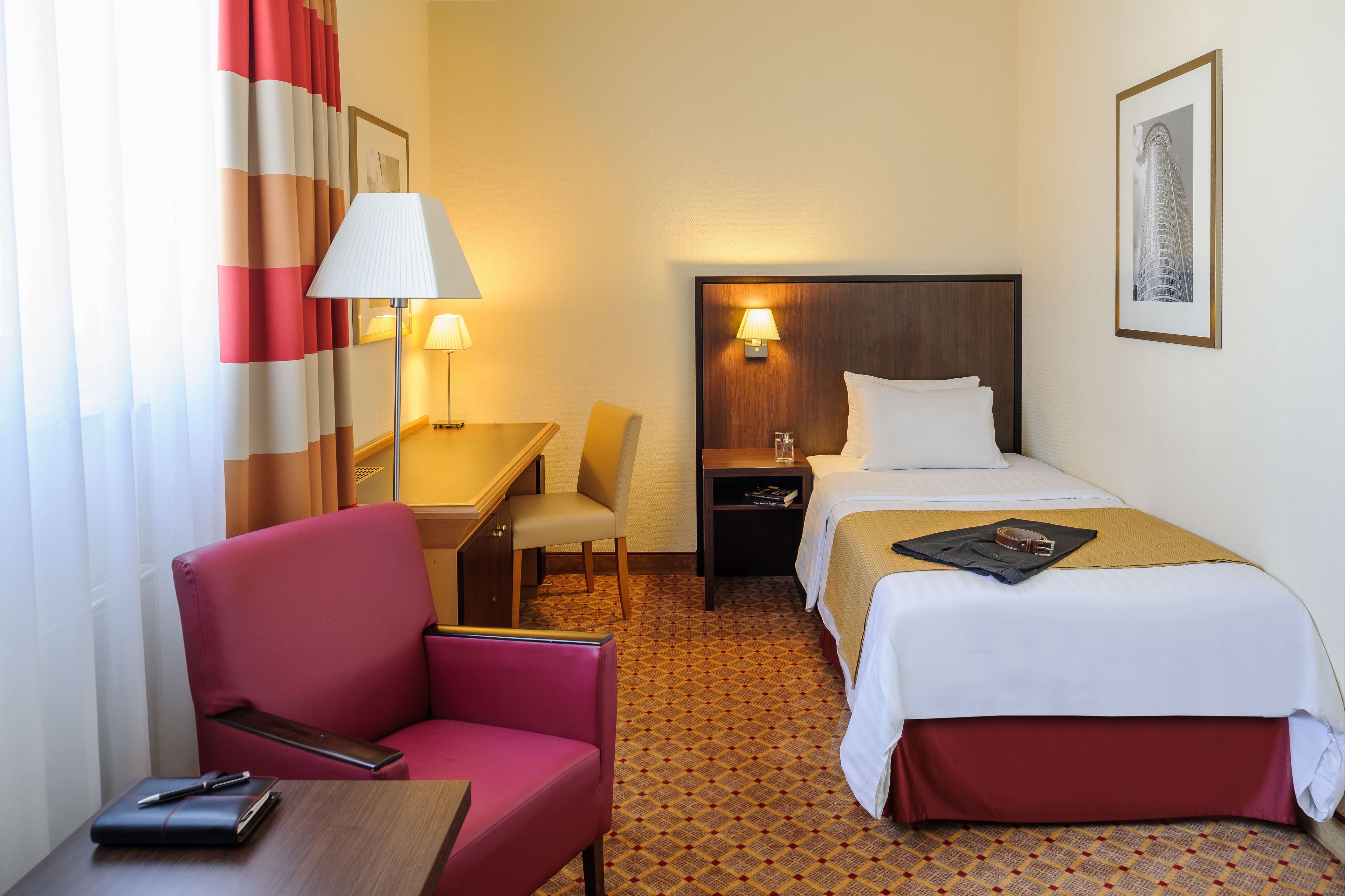 Habitación de hotel con cama, escritorio con silla, mesita de noche, lámpara y un cuadro en la pared.