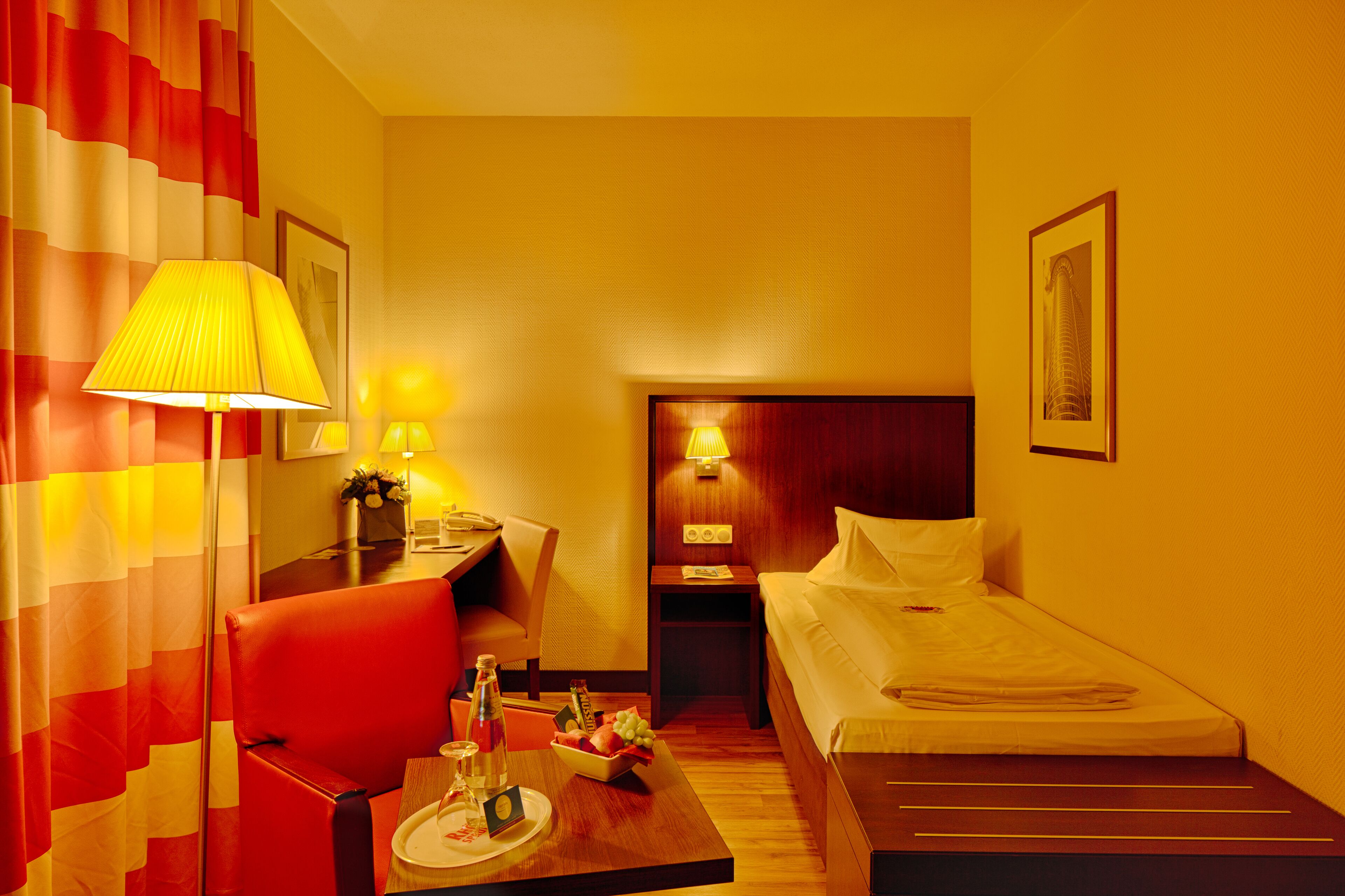 Habitación de hotel con cama, escritorio con lámpara, silla, mesita de noche con lámpara y un cuadro en la pared.