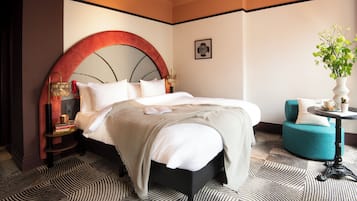 Quarto de hotel com uma cama grande, cabeceira redonda, uma mesinha com um vaso de flores e uma poltrona azul-esverdeada.