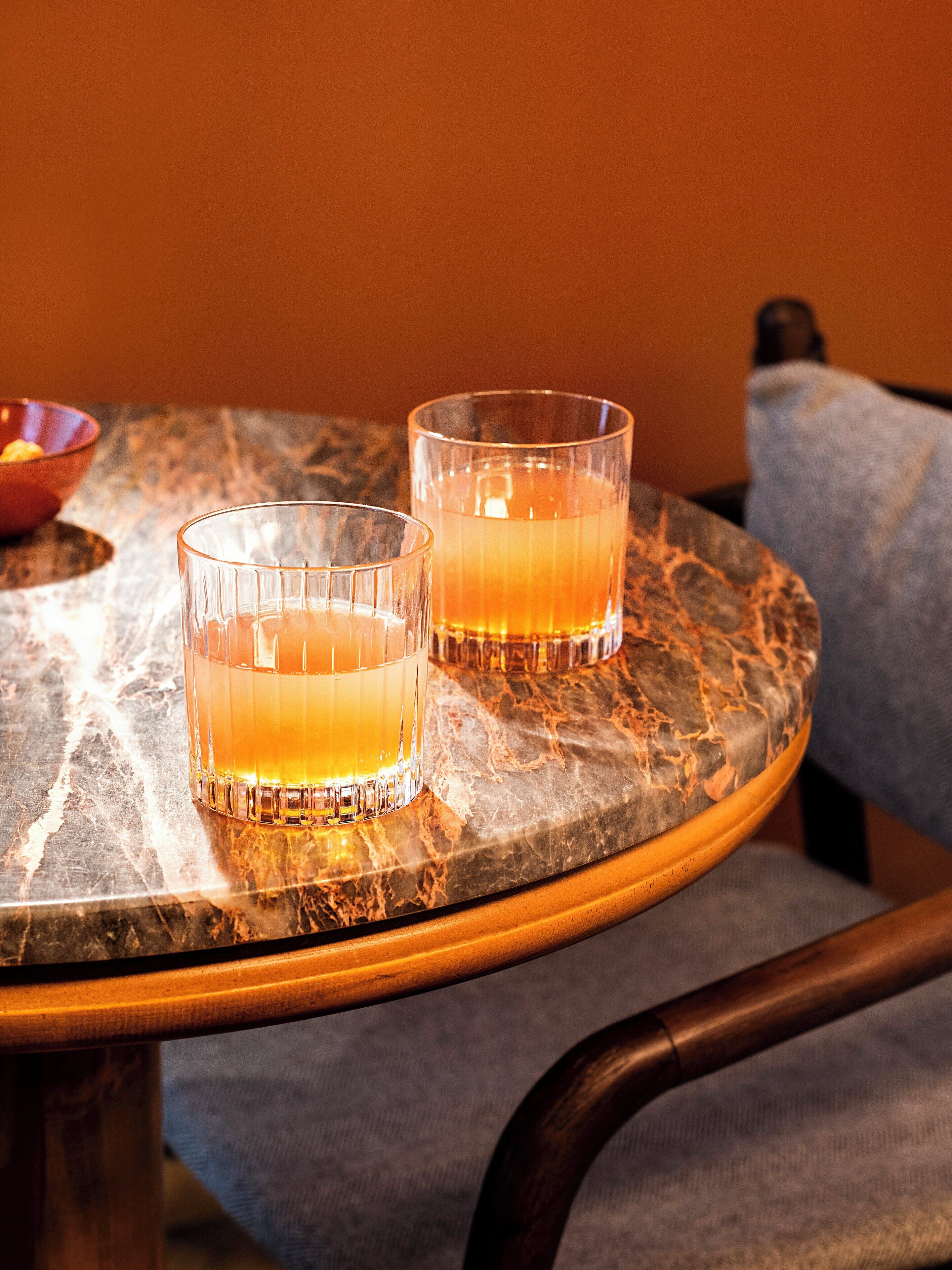 Two glasses of amber liquid on a marble table with a wooden chair.