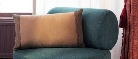 A teal armchair with two cushions, positioned in front of a window with sheer curtains.