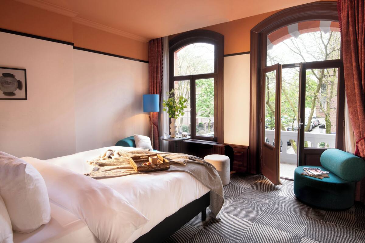 A hotel room with a large bed, a side table with a lamp, a window with curtains, and a view of the outside.