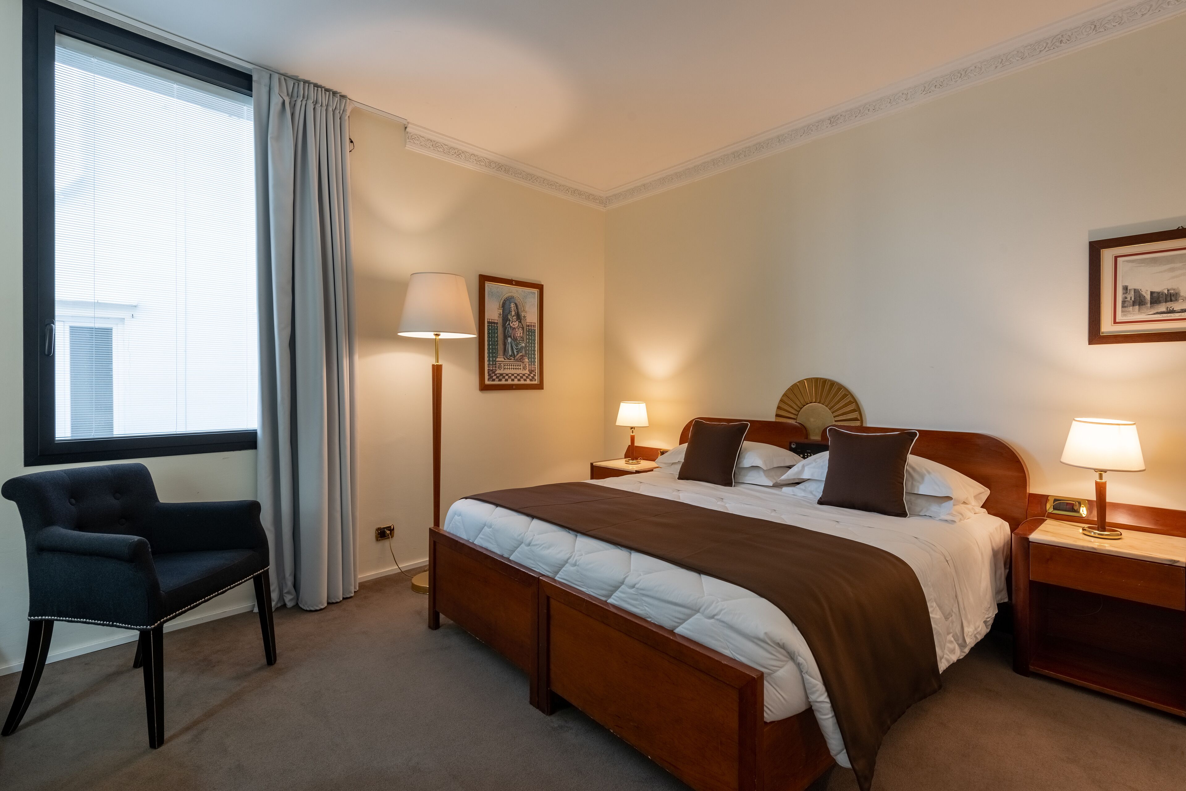 A hotel room with a large bed, a chair, two bedside tables with lamps, a window with curtains, and a framed picture on the wall.