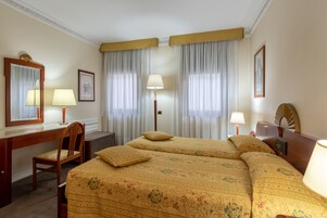 A hotel room with two beds, a desk, a chair, and a large window with curtains.