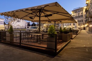 An outdoor café with tables and chairs under a large umbrella, surrounded by potted plants and a stone pathway.