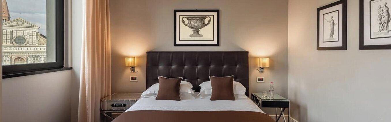 A hotel room with a large bed, two bedside lamps, a framed picture above the bed, and a window with a view of a building.