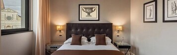 A hotel room with a large bed, two bedside lamps, a framed picture above the bed, and a window with a view of a building.