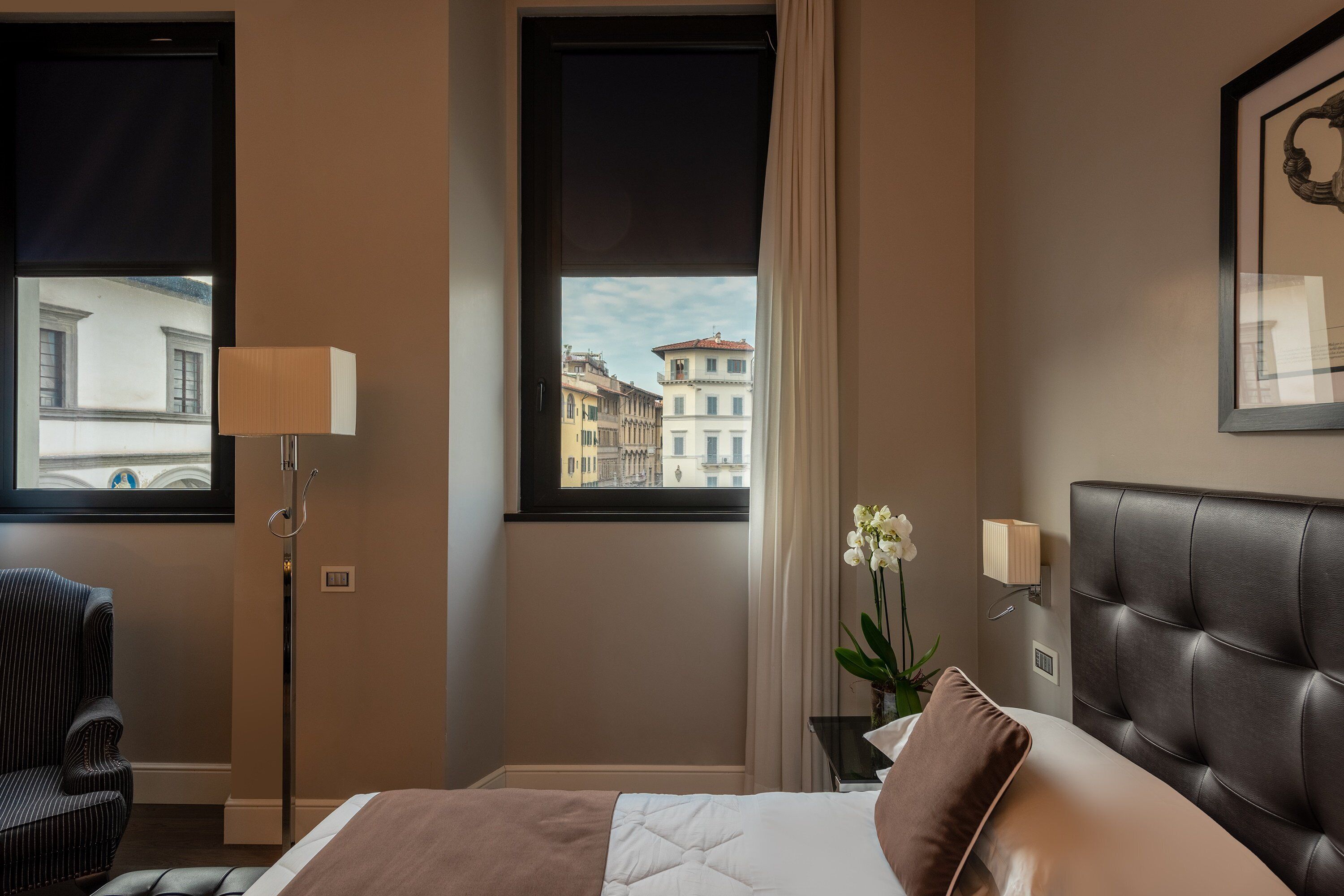 A hotel room with a bed, a chair, a nightstand with a lamp, and a window with a view of buildings.