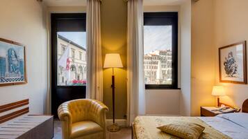 A hotel room with a bed, a chair, a floor lamp, and two windows with curtains.