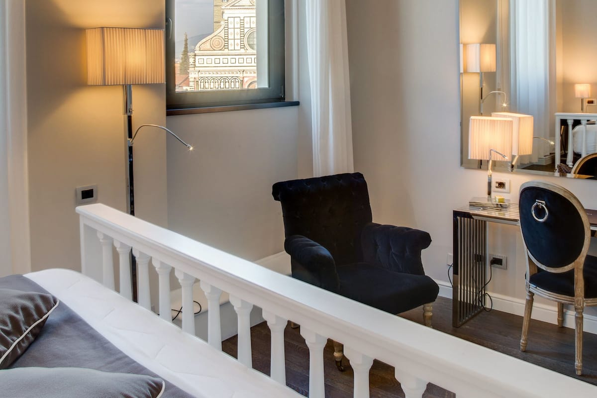 A hotel room with a bed, a chair, a desk with a lamp, a window with a view of a building, and a balcony railing.