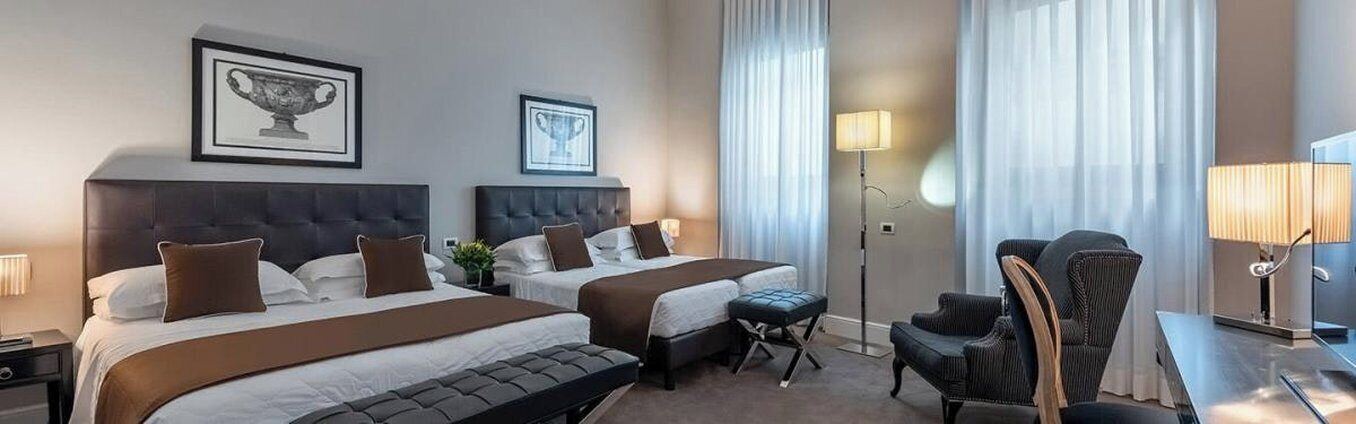 A hotel room with two beds, a desk, a chair, and a lamp.