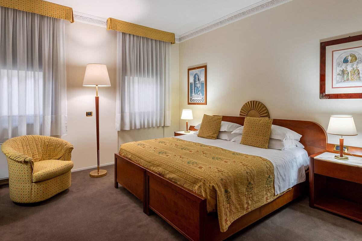 A hotel room with a large bed, a wooden nightstand, a floor lamp, a chair, and two windows with curtains.
