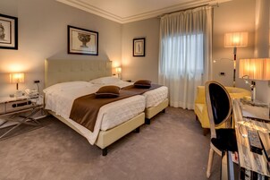 Una habitación de hotel con dos camas, un escritorio con silla, una lámpara y obras de arte enmarcadas en las paredes.
