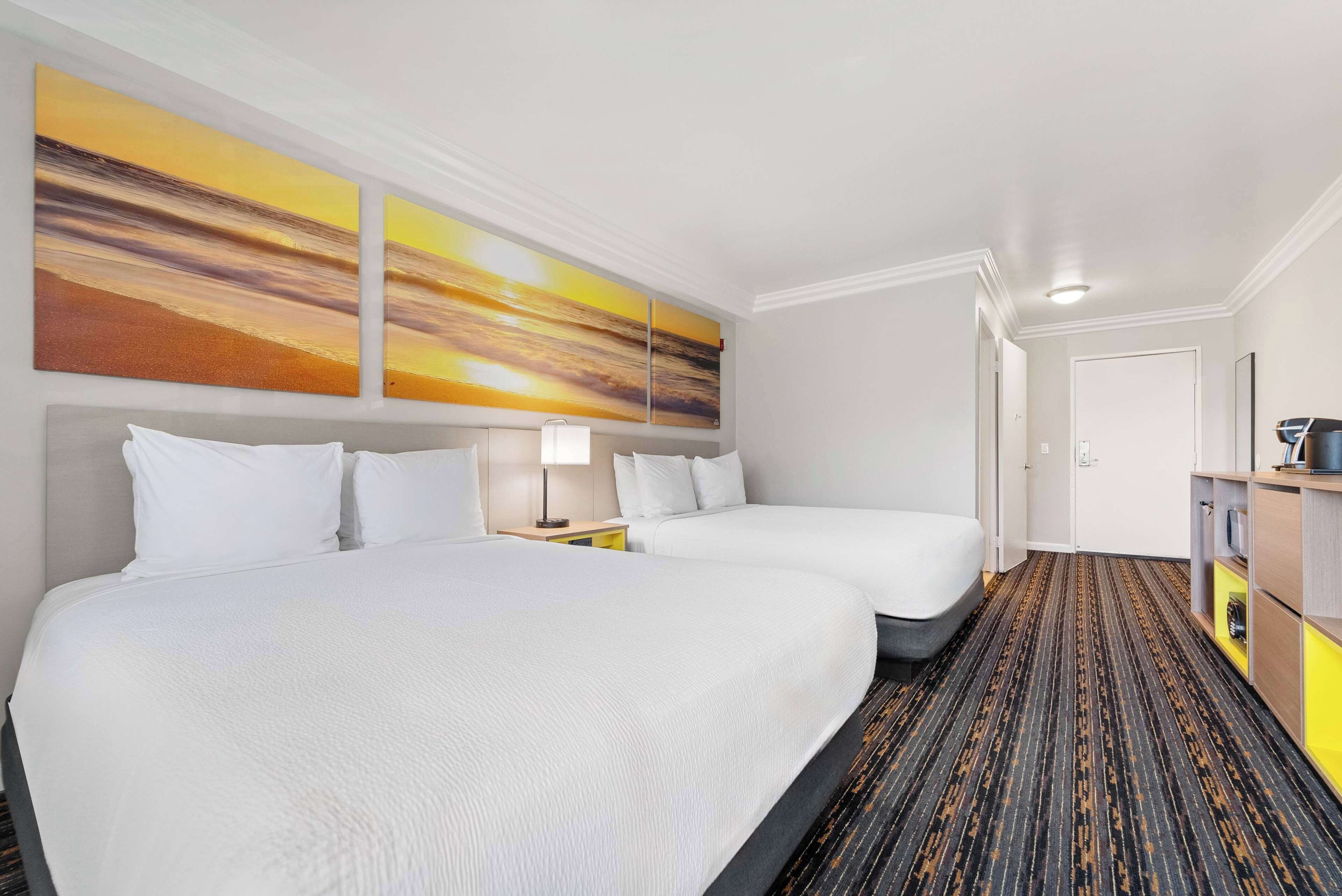 A hotel room with two beds, a large painting on the wall, a carpeted floor, and a small desk with a lamp.