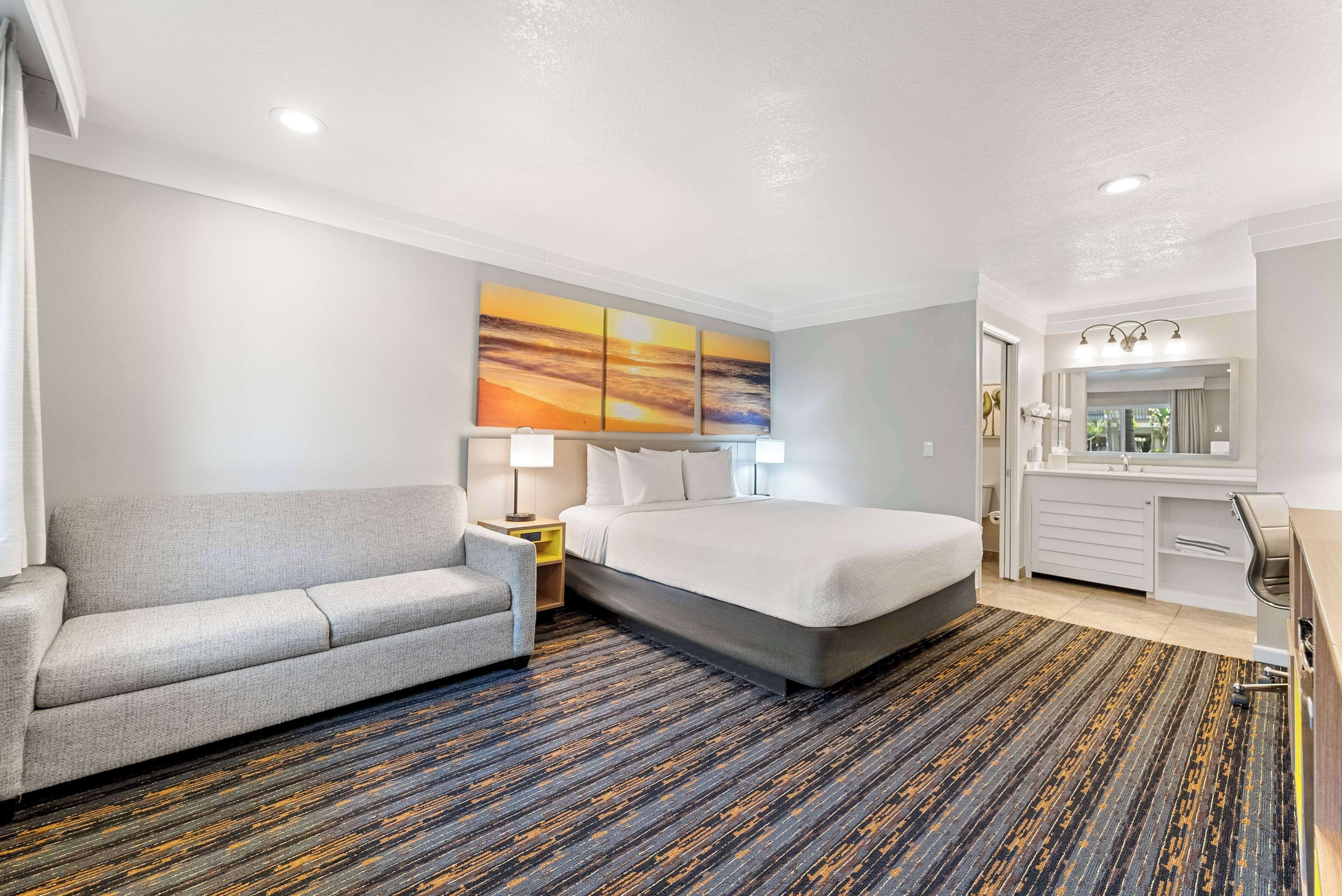 A hotel room with a sofa, a bed, a desk, a mirror, and a painting on the wall.
