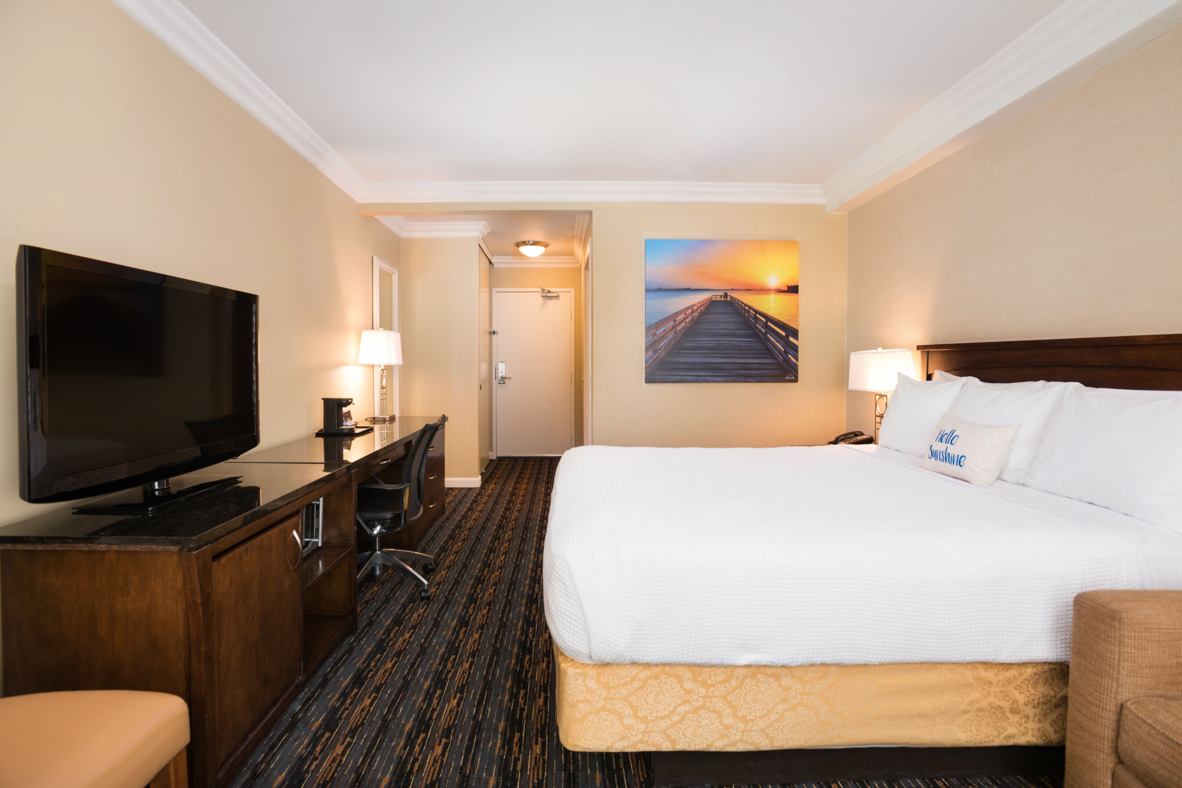 A hotel room with a large bed, a flat-screen TV, a desk with a chair, and a painting of a sunset over a pier.