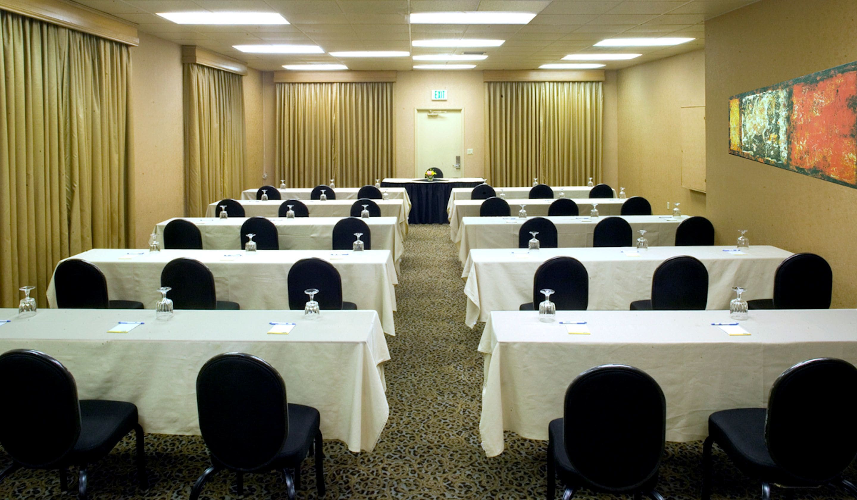 A conference room with rows of tables and chairs, an exit sign, and a painting on the wall.
