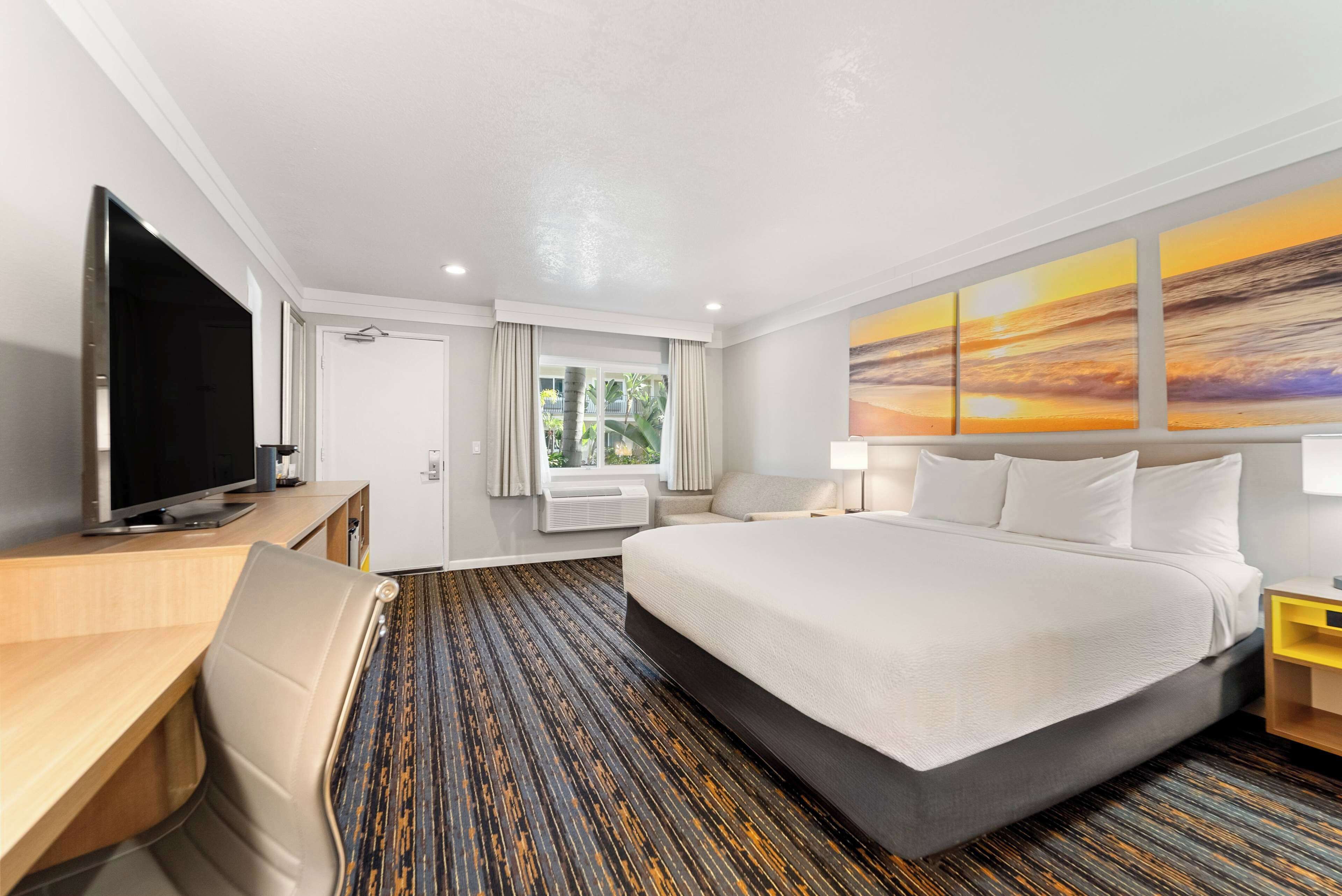 A hotel room with a large bed, a desk with a flat-screen TV, a chair, a window with curtains, an air conditioning unit, and a wall with a sunset painting.