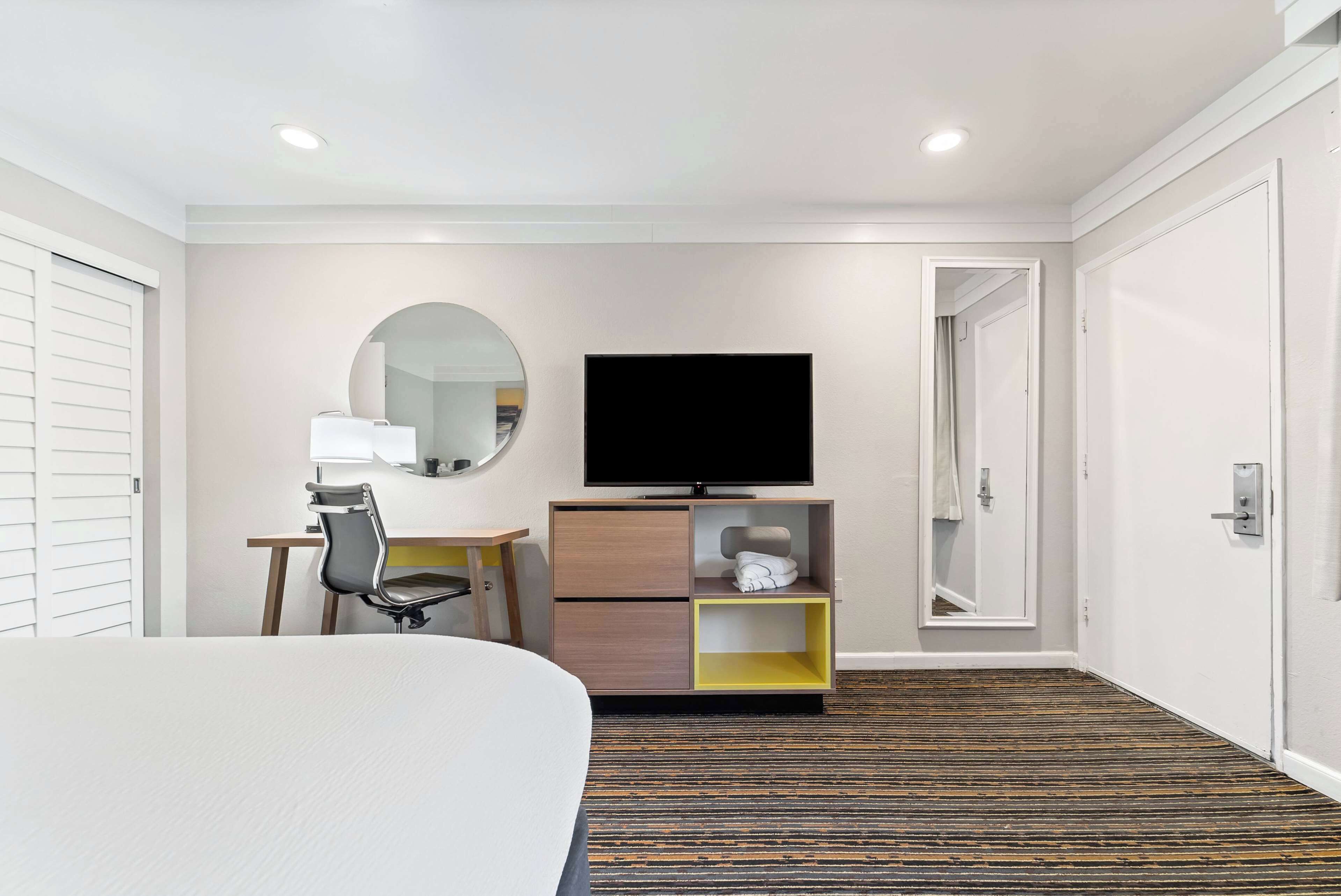 A hotel room with a bed, a desk with a chair, a television, a mirror, and a wardrobe.