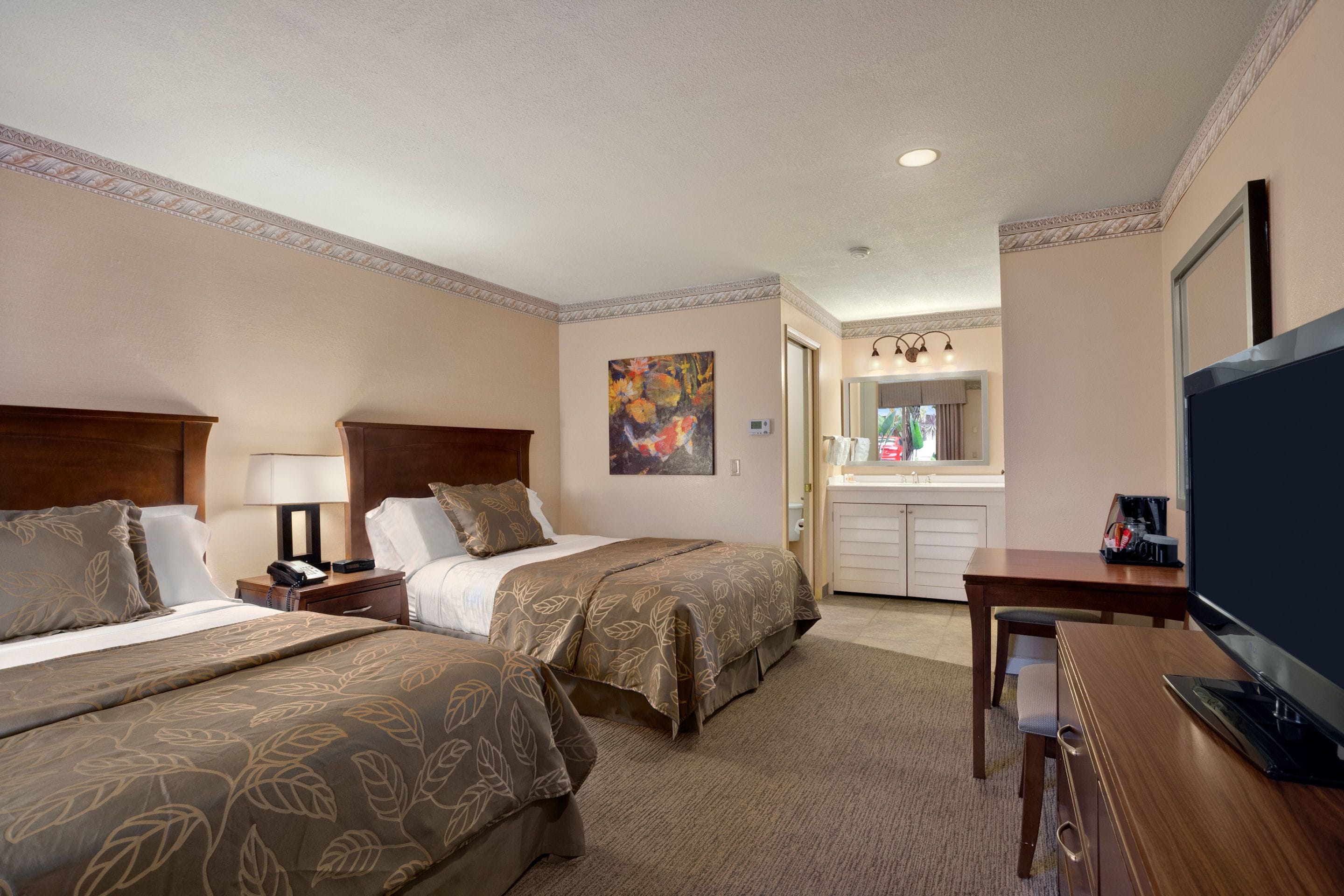 A hotel room with two beds, a desk, a TV, a painting, and a bathroom visible through an open door.