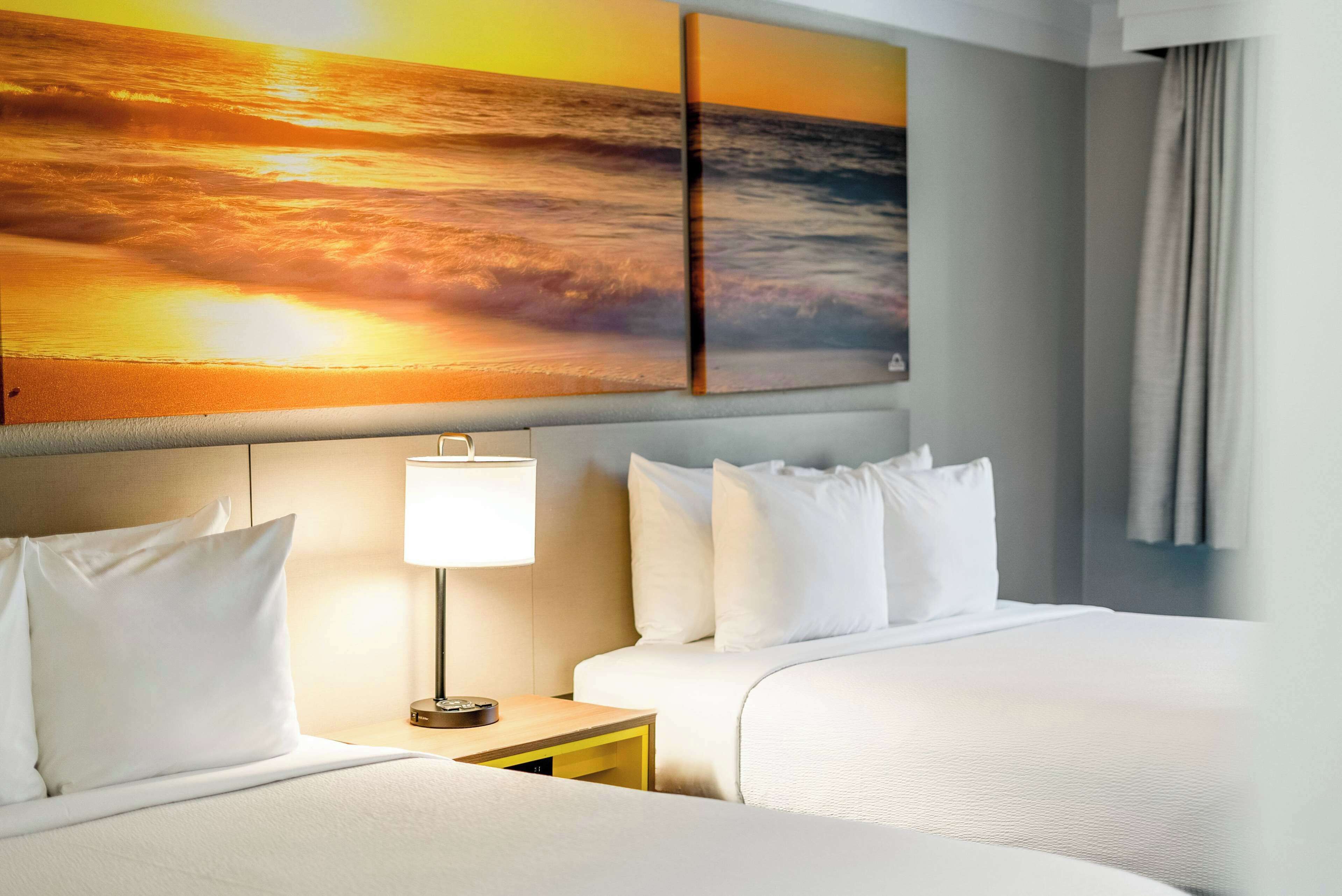 Quarto de hotel com duas camas, uma mesinha de cabeceira, uma luminária e uma grande pintura de pôr do sol acima das camas.