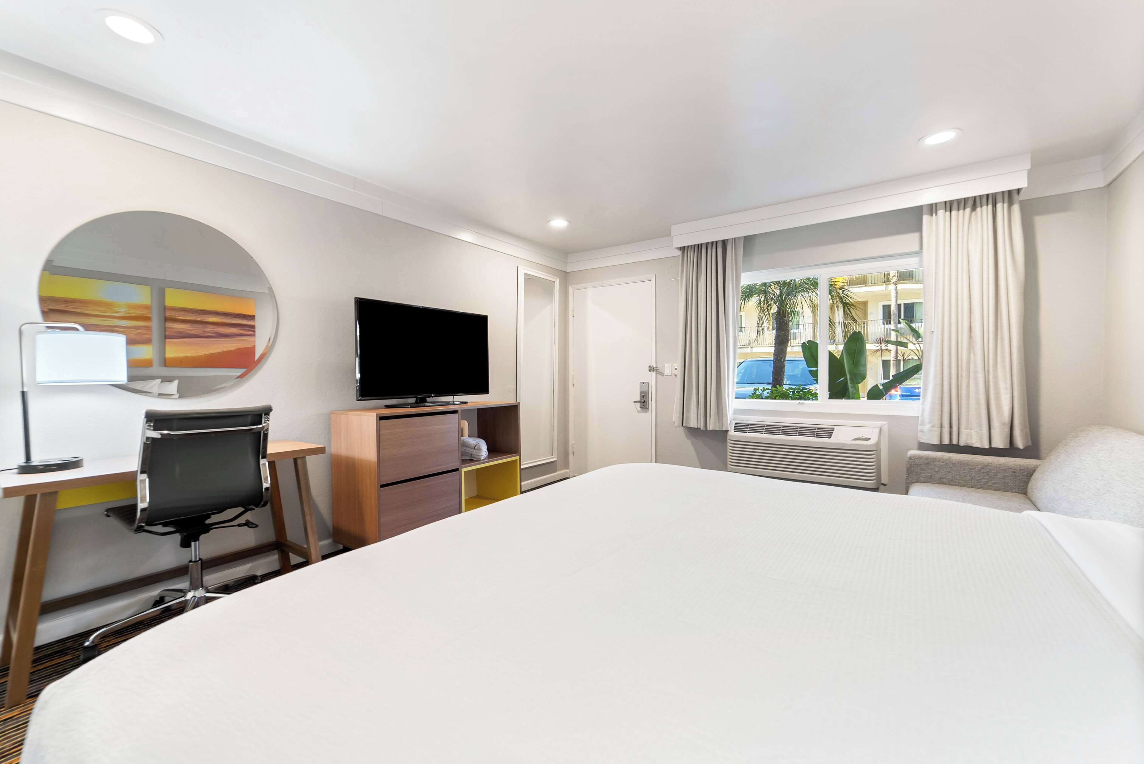 A hotel room with a large bed, a desk with a chair, a flat-screen TV, a wardrobe, and a window with curtains.
