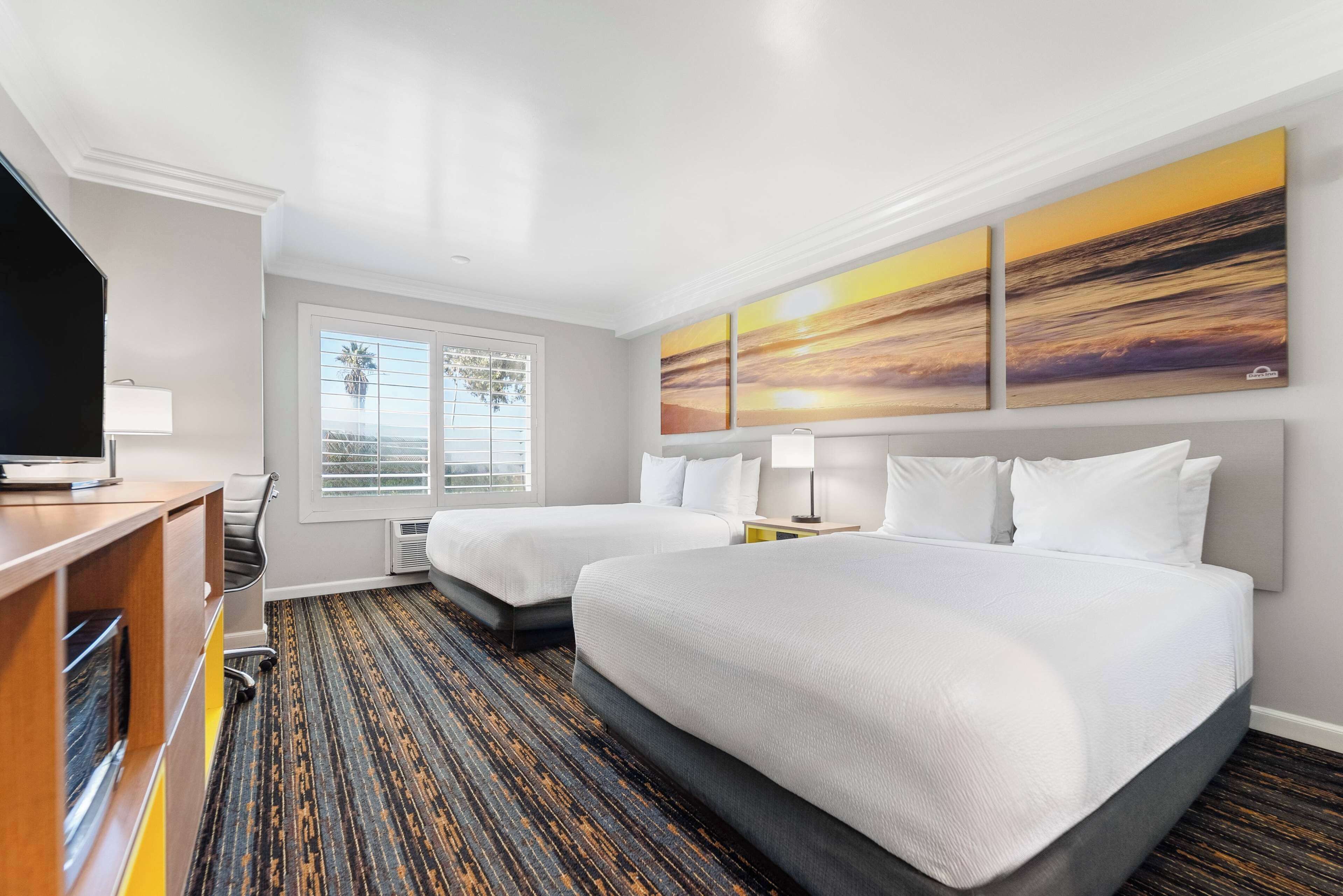 A hotel room with two beds, a desk with a TV, a window with a view, and a painting on the wall.