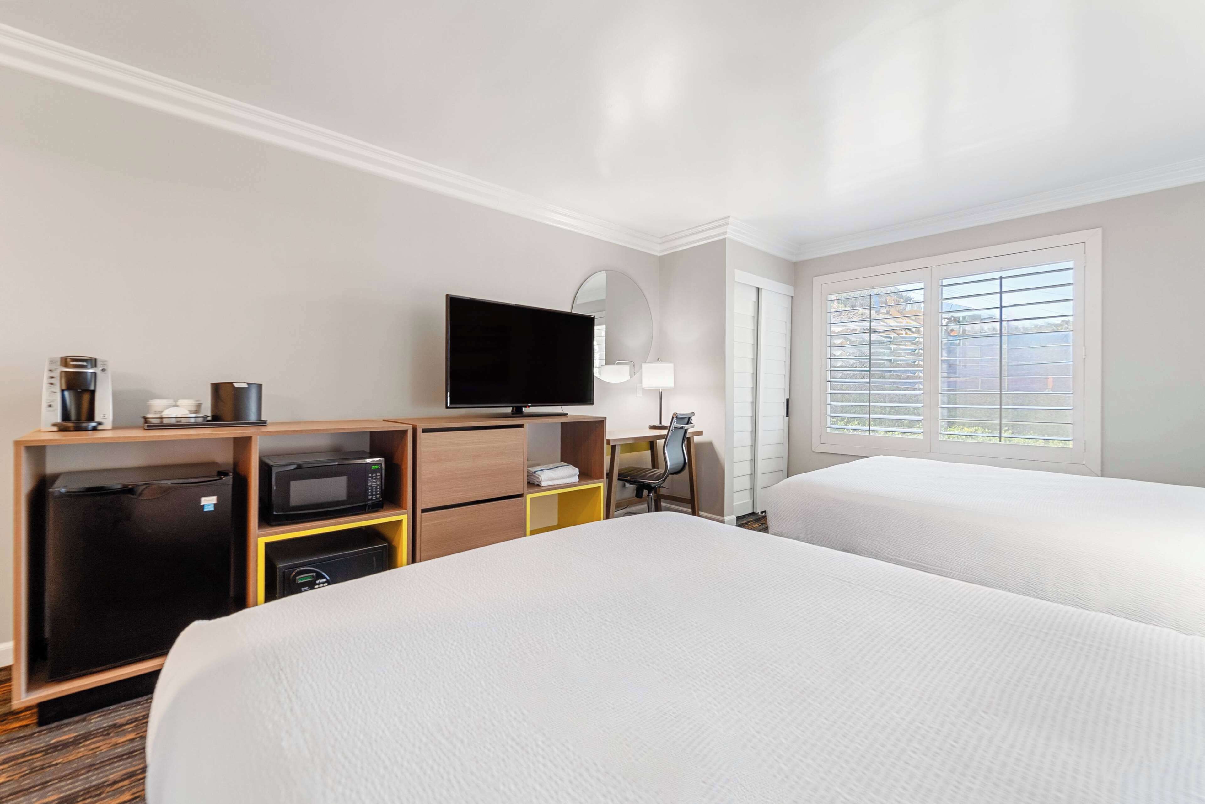 A hotel room with two beds, a television, a desk with a chair, a microwave, a refrigerator, and a window with blinds.
