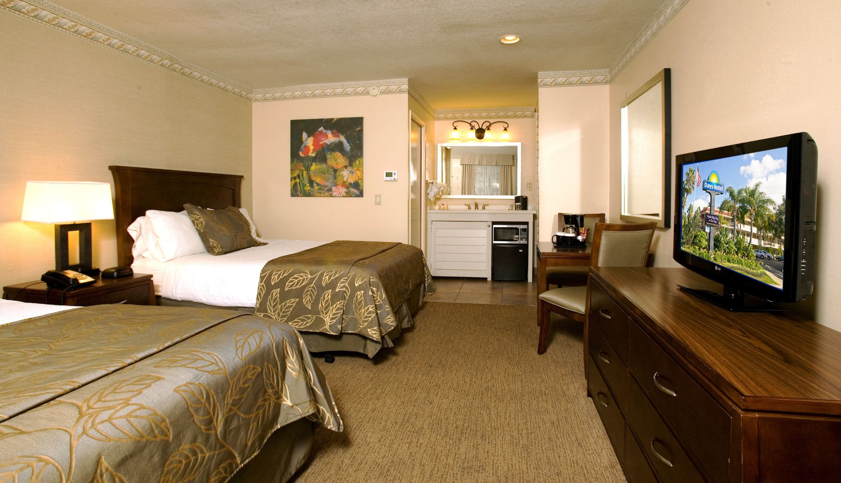 A hotel room with two beds, a desk with a television, a kitchenette, and a bathroom visible through an open door.
