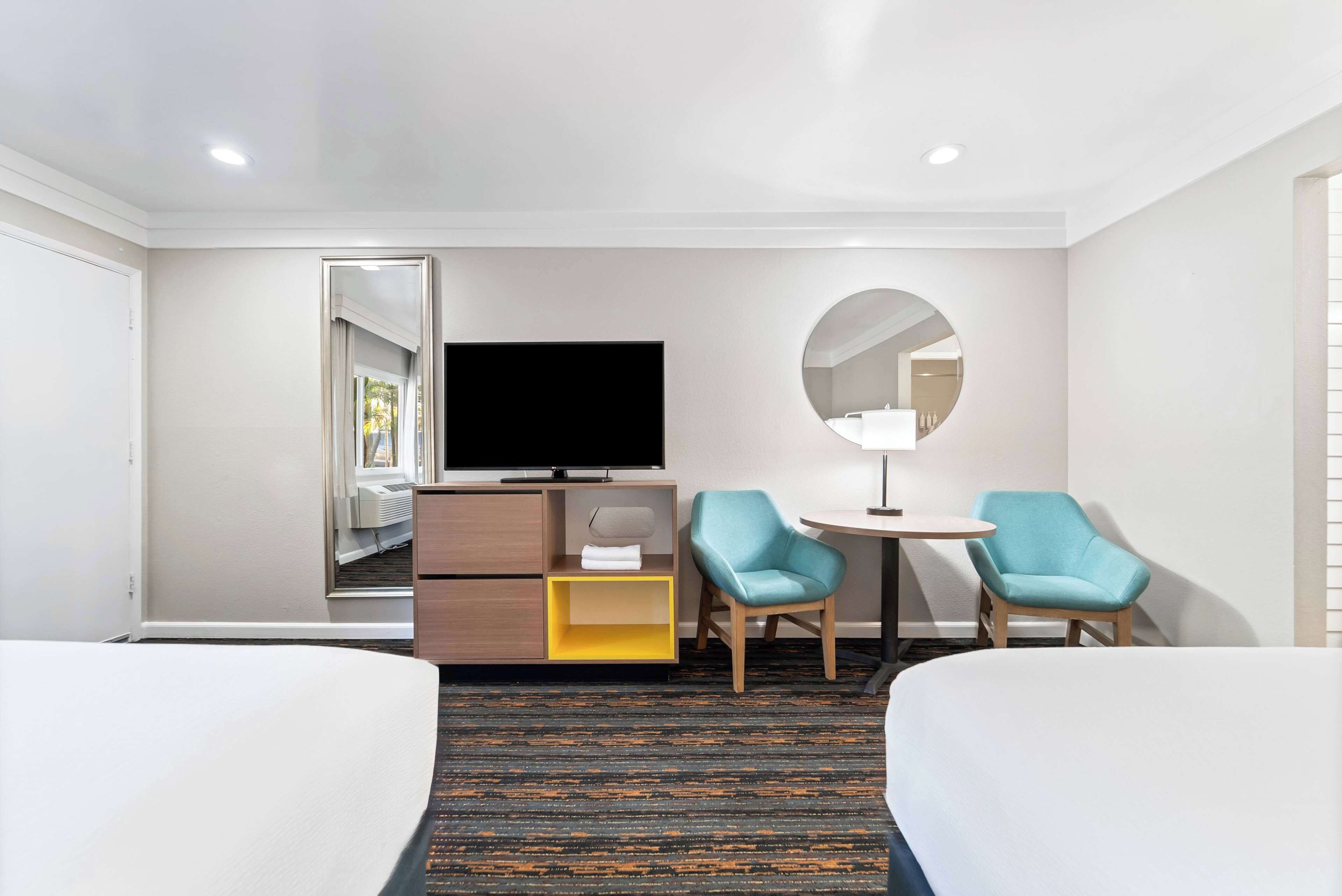 A hotel room with a bed, a desk with a TV, two chairs, a small table, and a mirror.
