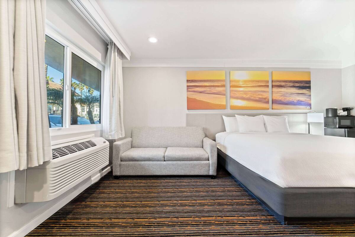 Ein Hotelzimmer mit Bett, Sofa, Stuhl, einem Fenster mit Vorhängen und einer Wand mit drei Sonnenuntergangsbildern.